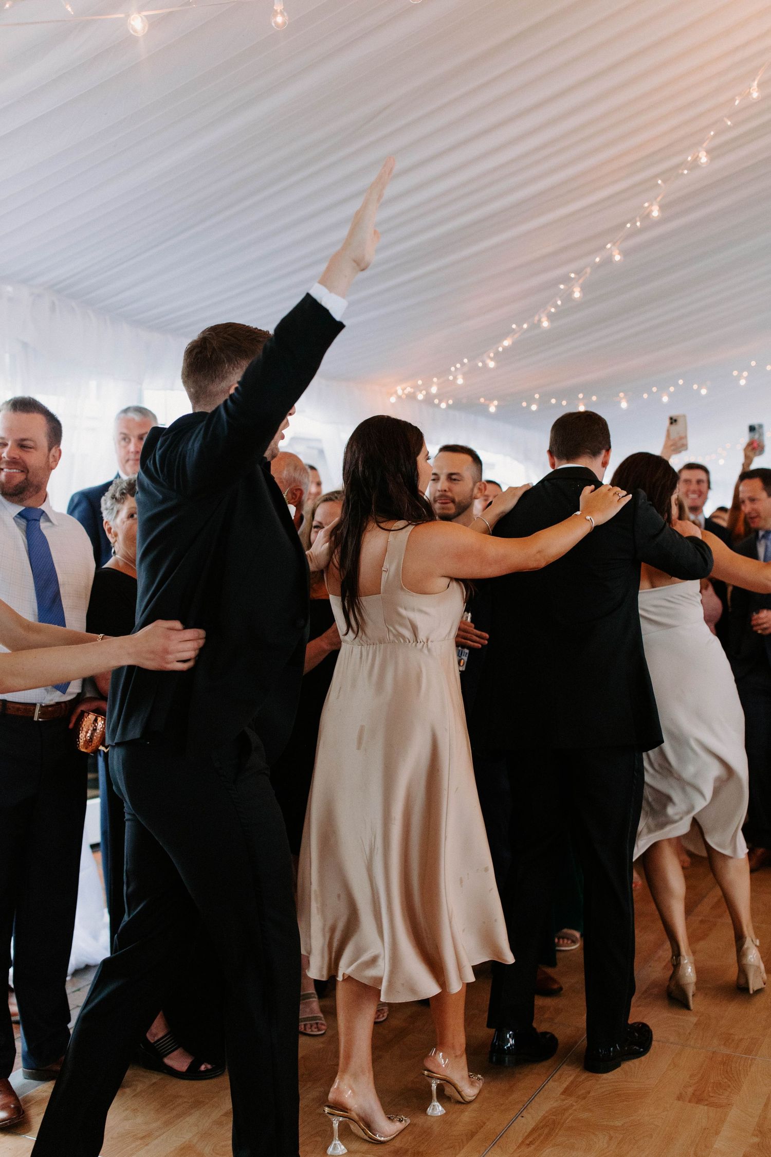 Wedding guests dancing together in a white tent with string lights overhead.