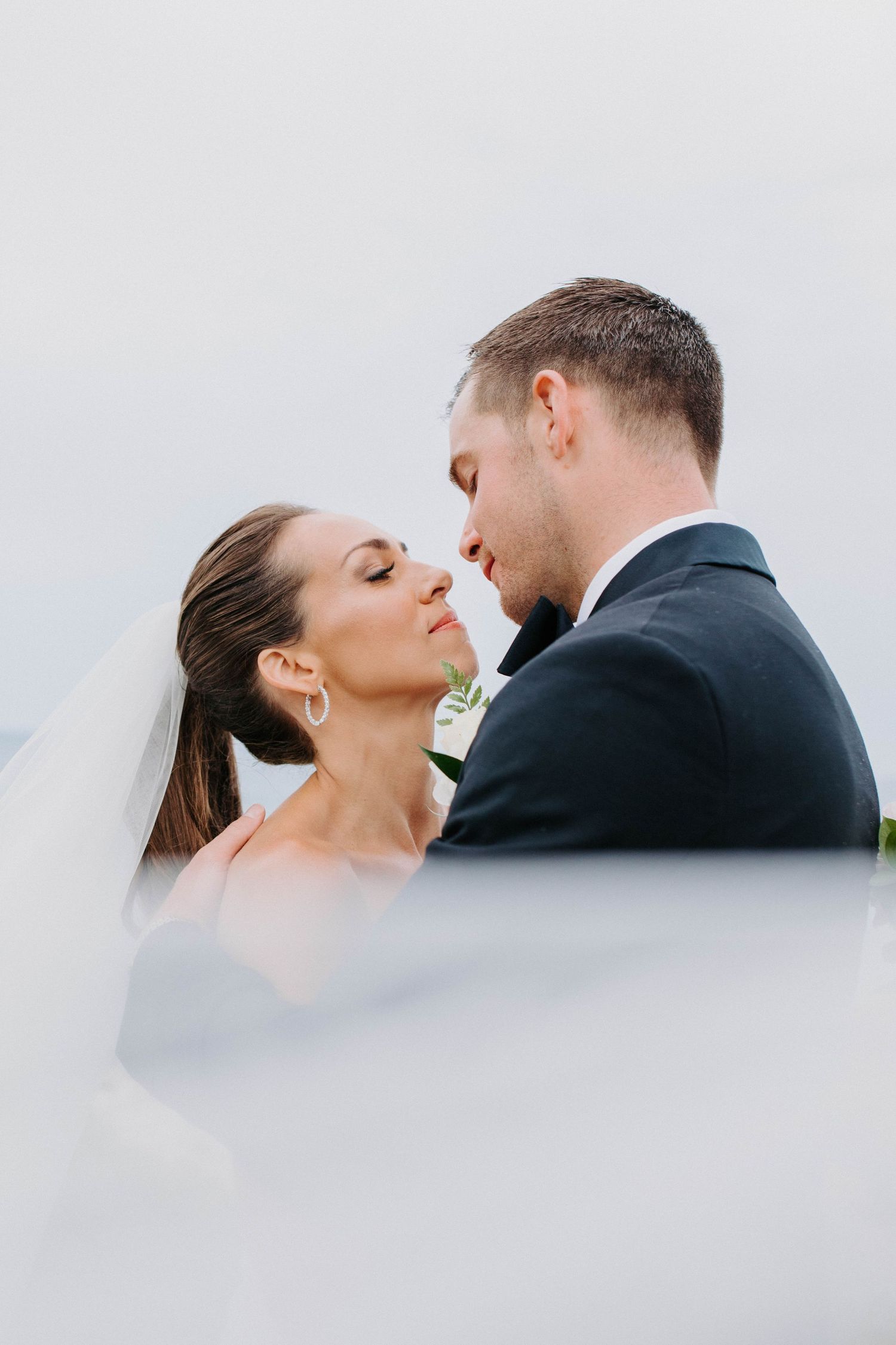 Intimate wedding portrait with soft lighting and flowing veil against sky.