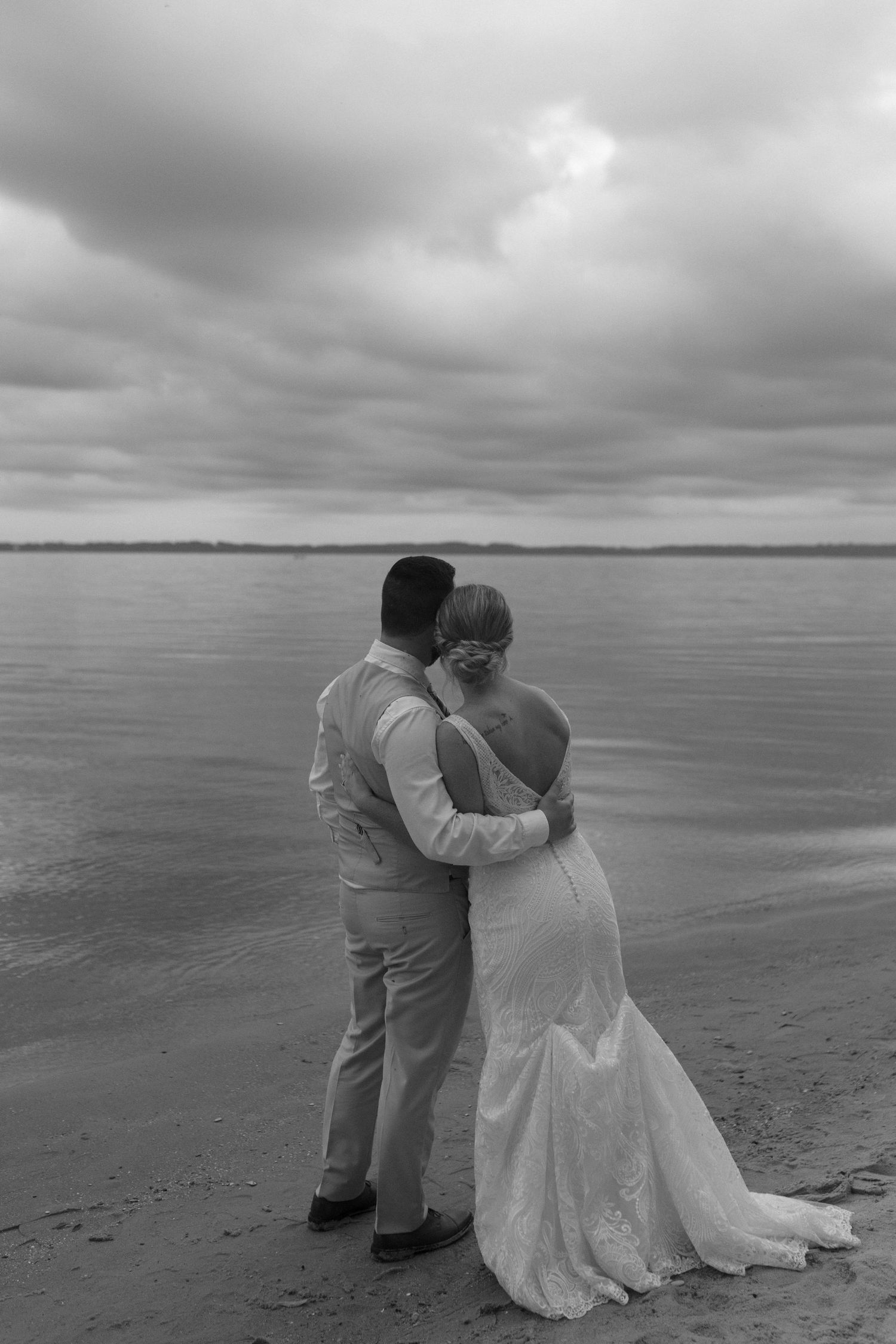A couple in wedding attire shares an intimate moment on a beach under moody grey skies.