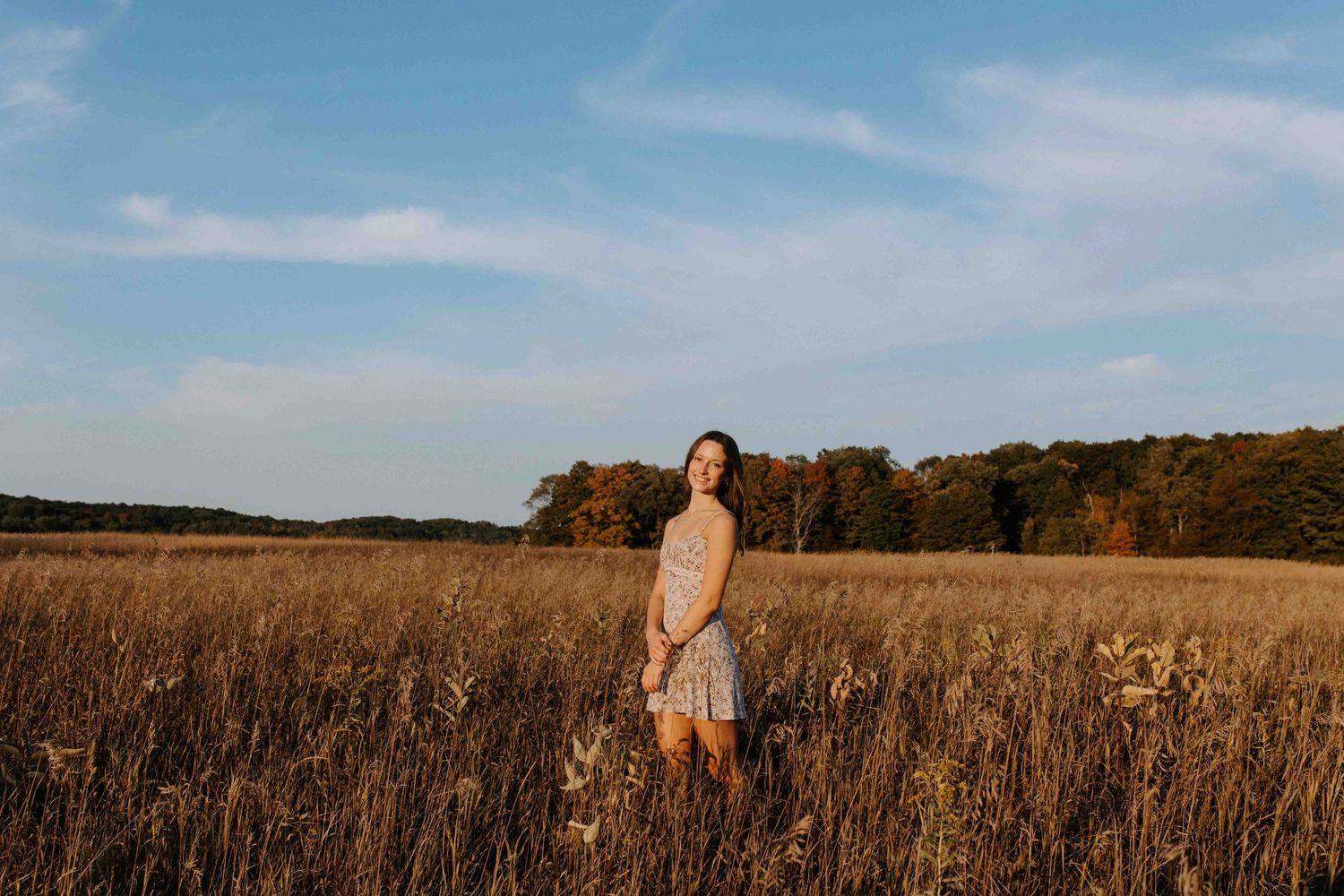 A person stands in a golden field at sunset with autumn trees in the background.