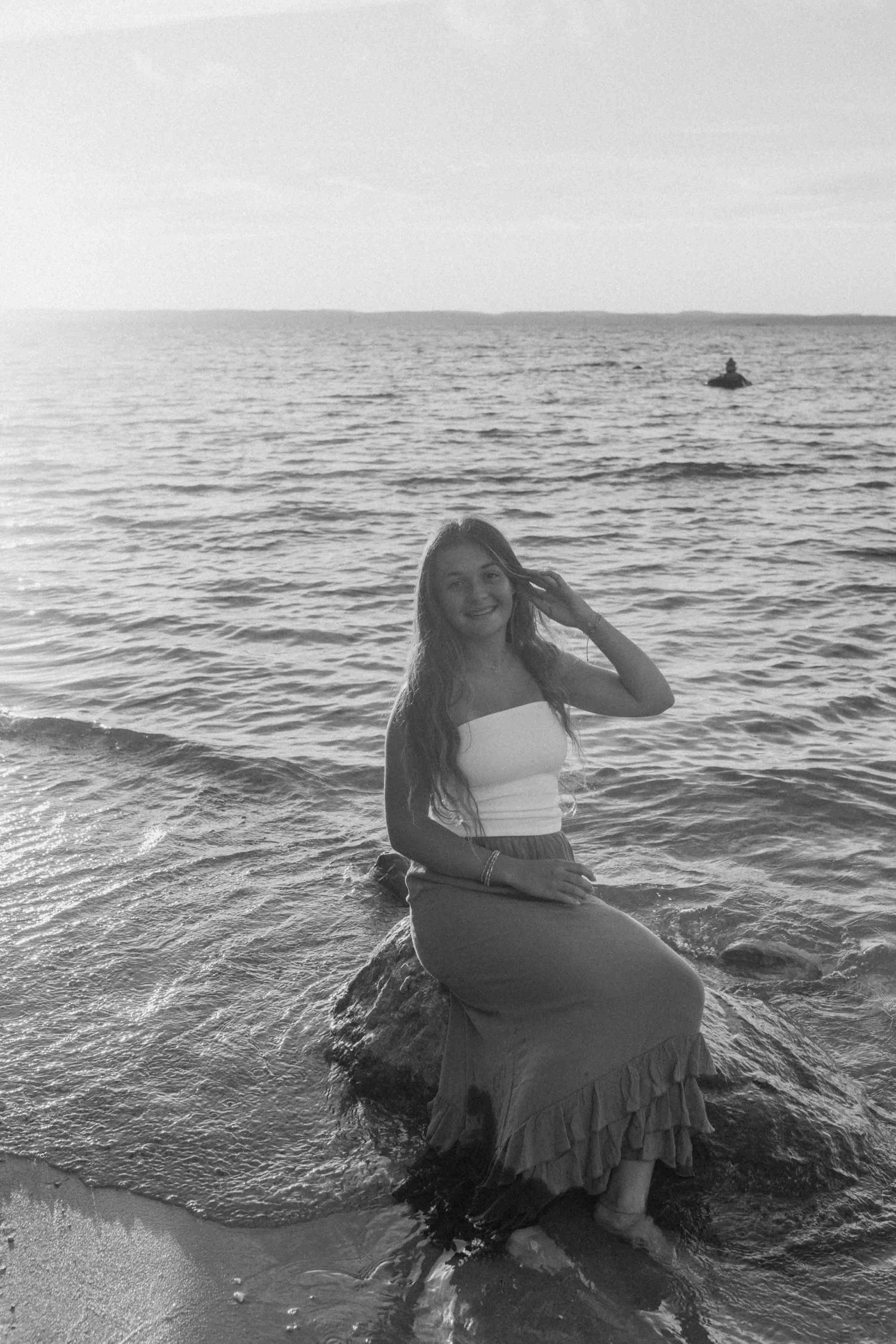 Black and white portrait of someone in a ruffled skirt sitting on coastal rocks by the ocean.
