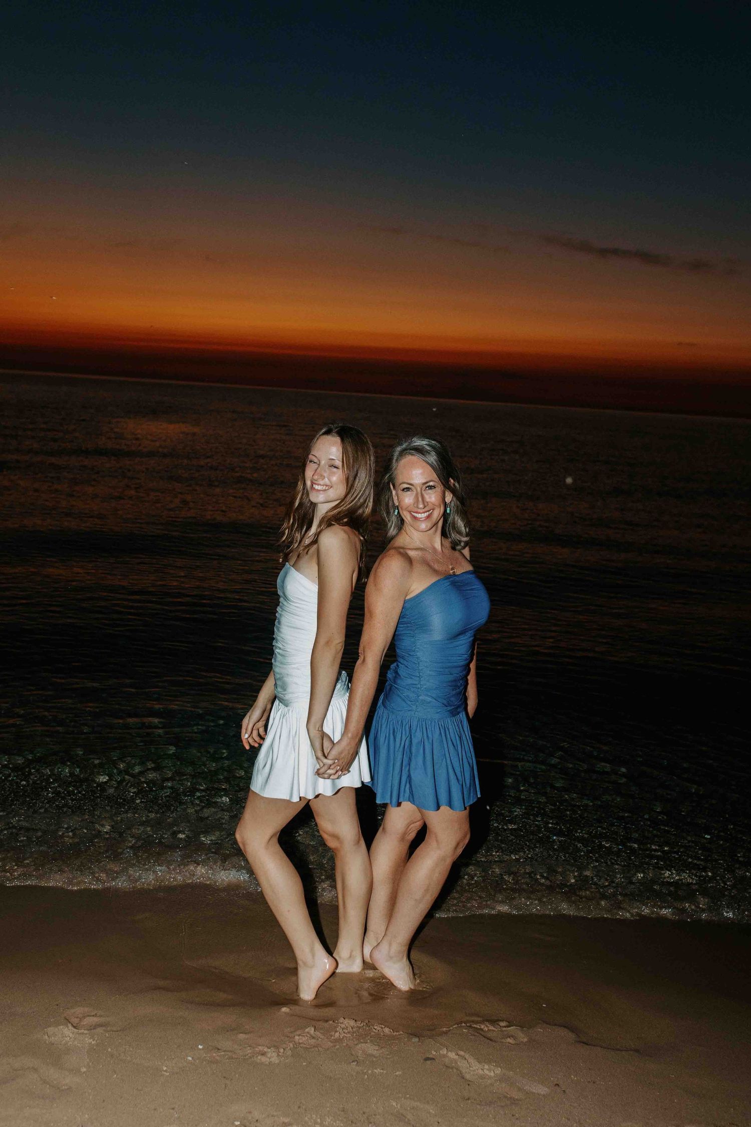 Two friends in white and blue dresses share a moment on the beach at dusk.