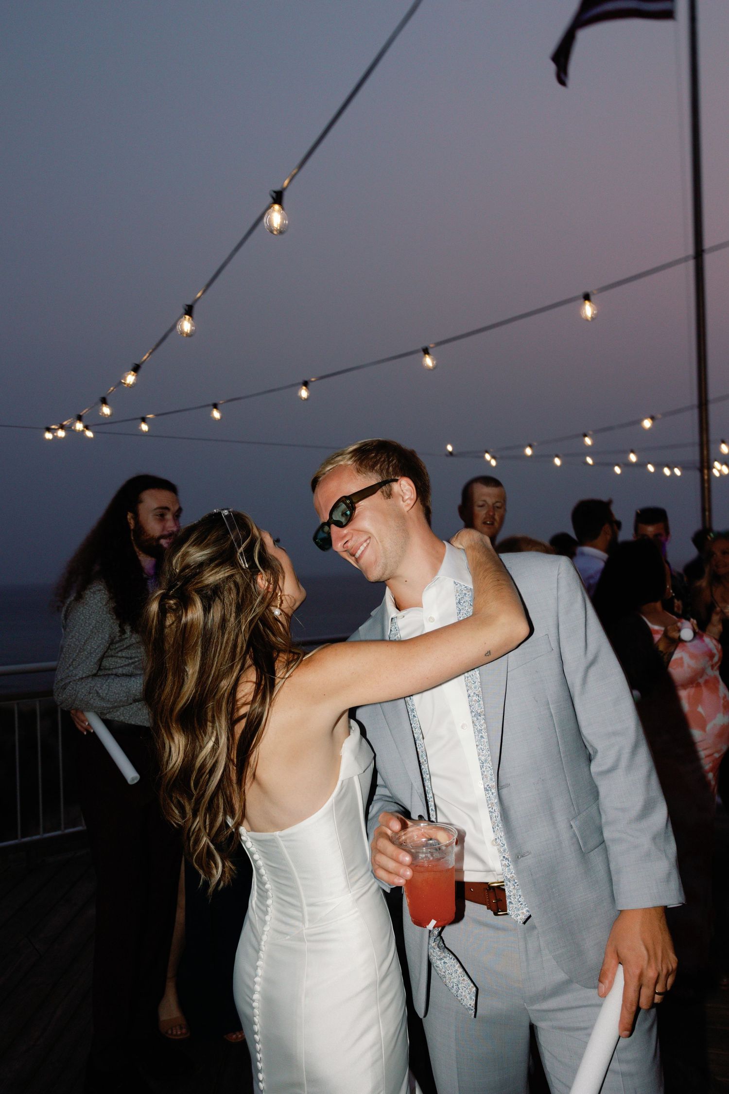 A couple dances together under string lights at an outdoor evening reception.
