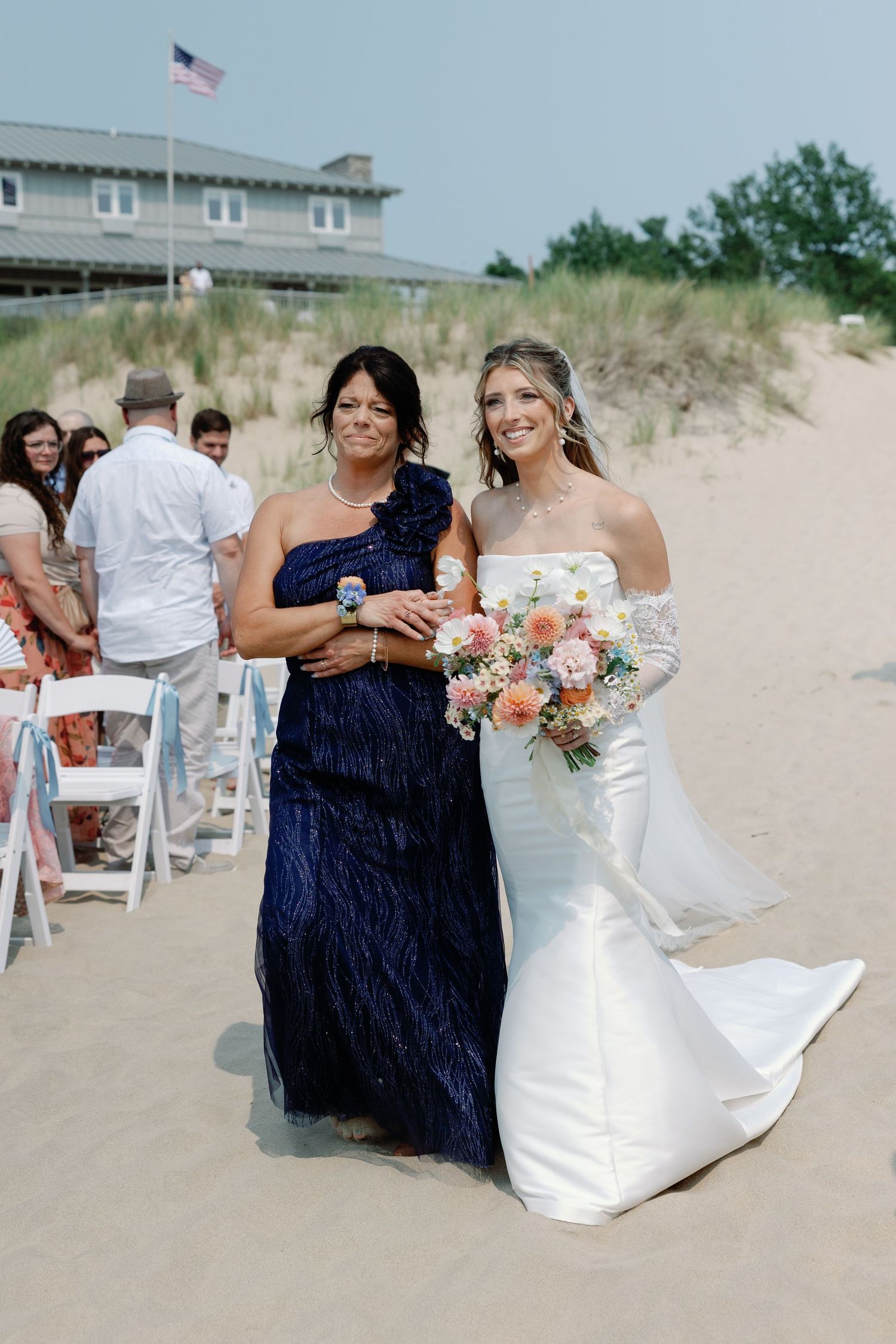 Wedding party on a sandy beach with white chairs and floral arrangements.