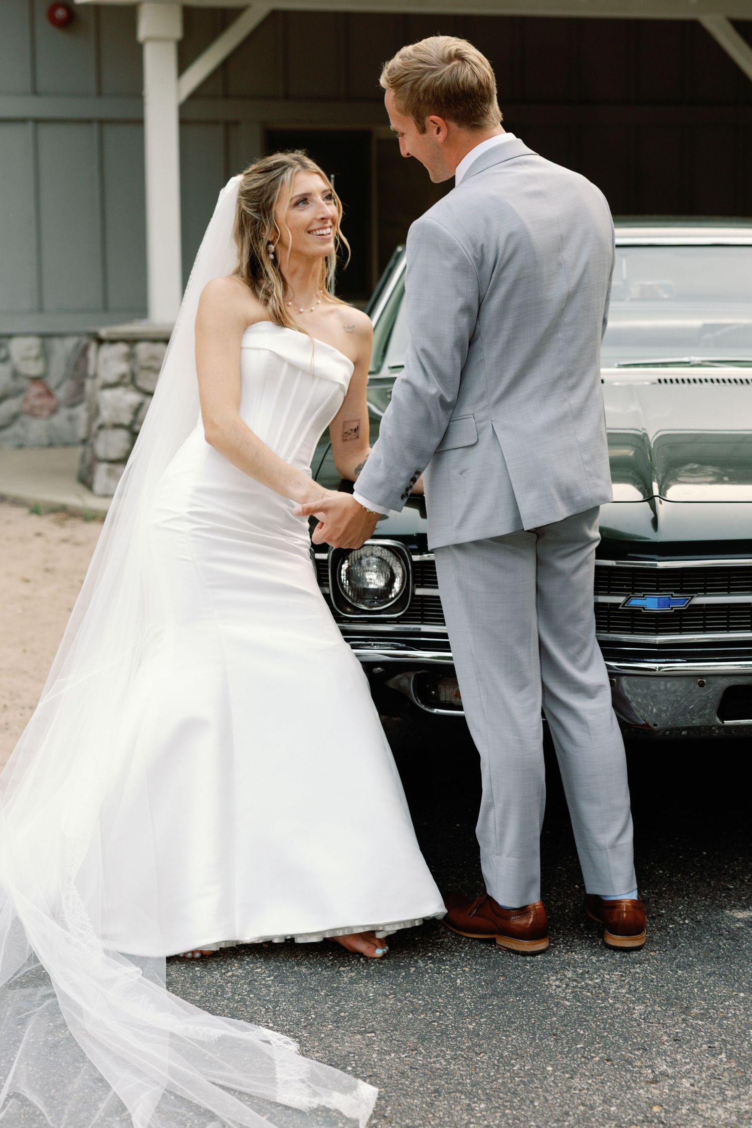 Bride in flowing white dress and groom in gray suit share playful moments near a vintage car.