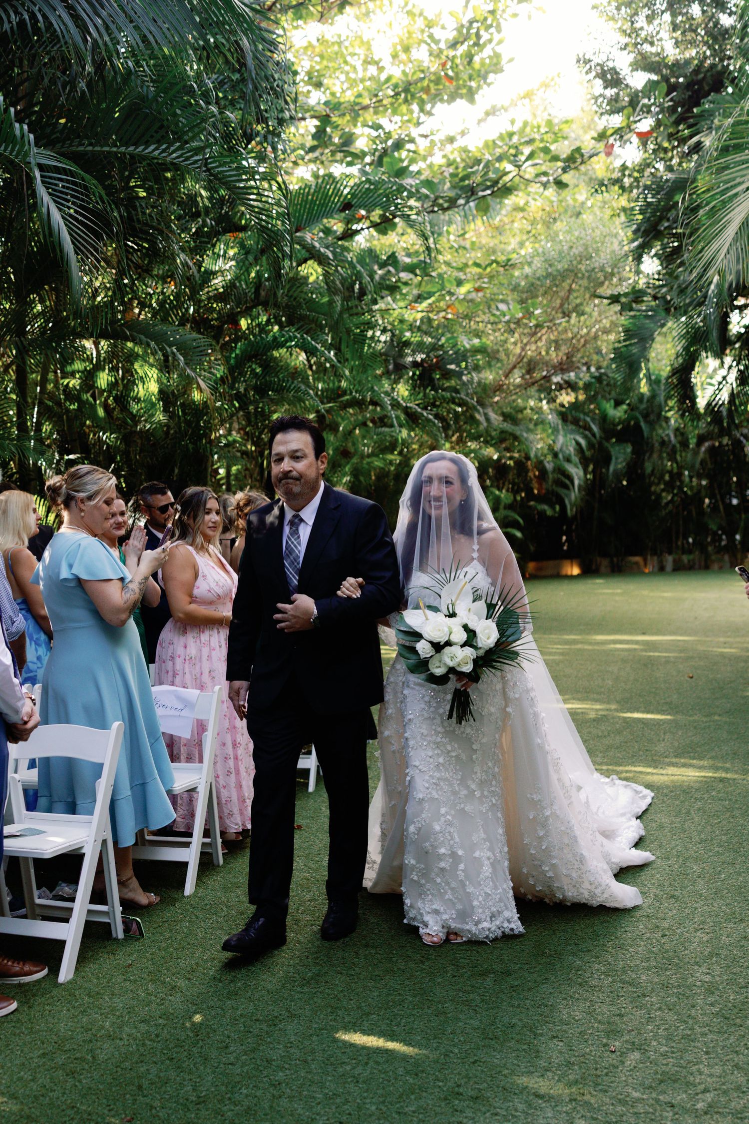 Outdoor garden wedding ceremony with guests seated on white chairs among tropical foliage.