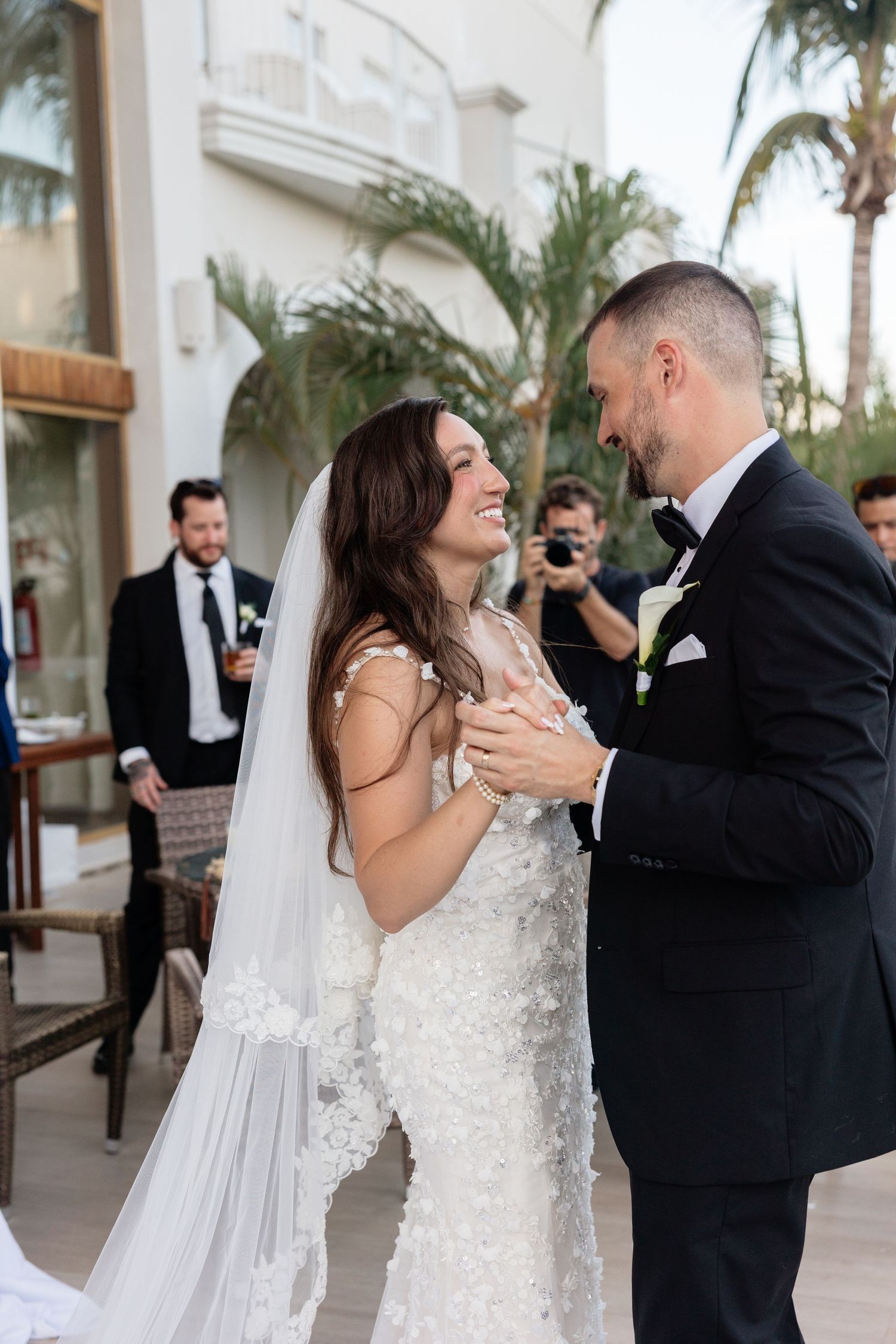 Couple dances on outdoor terrace with palm trees and white columns in background.