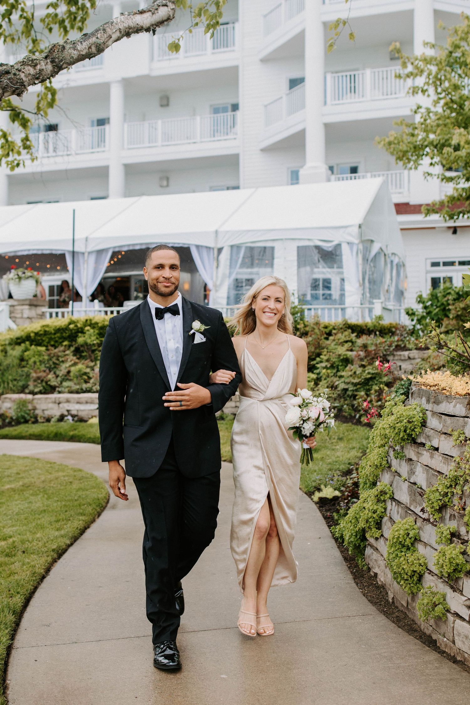Wedding party members walk arm in arm along a curved stone path at an elegant resort.