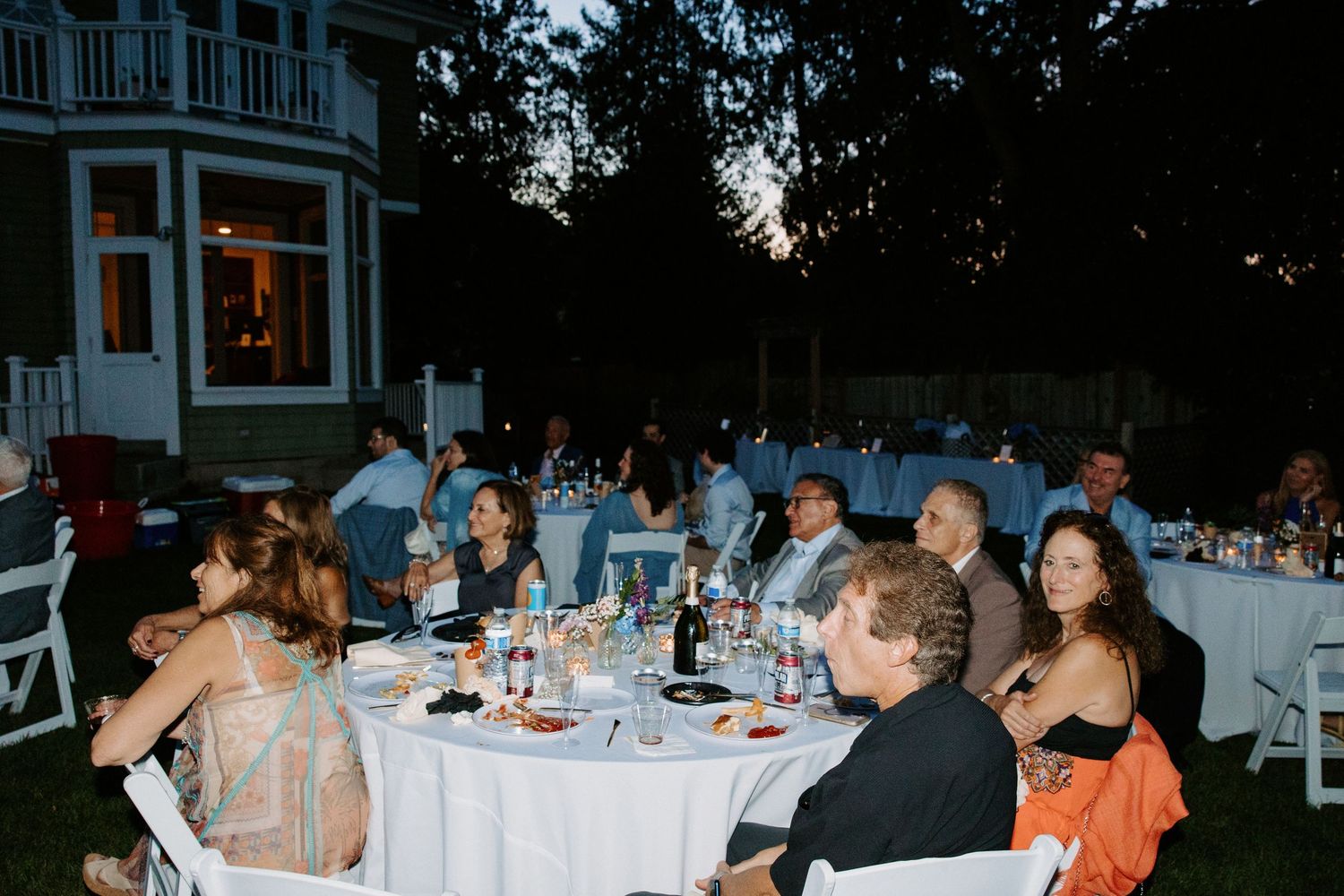 Long tables set for an outdoor wedding reception with guests seated under evening sky.