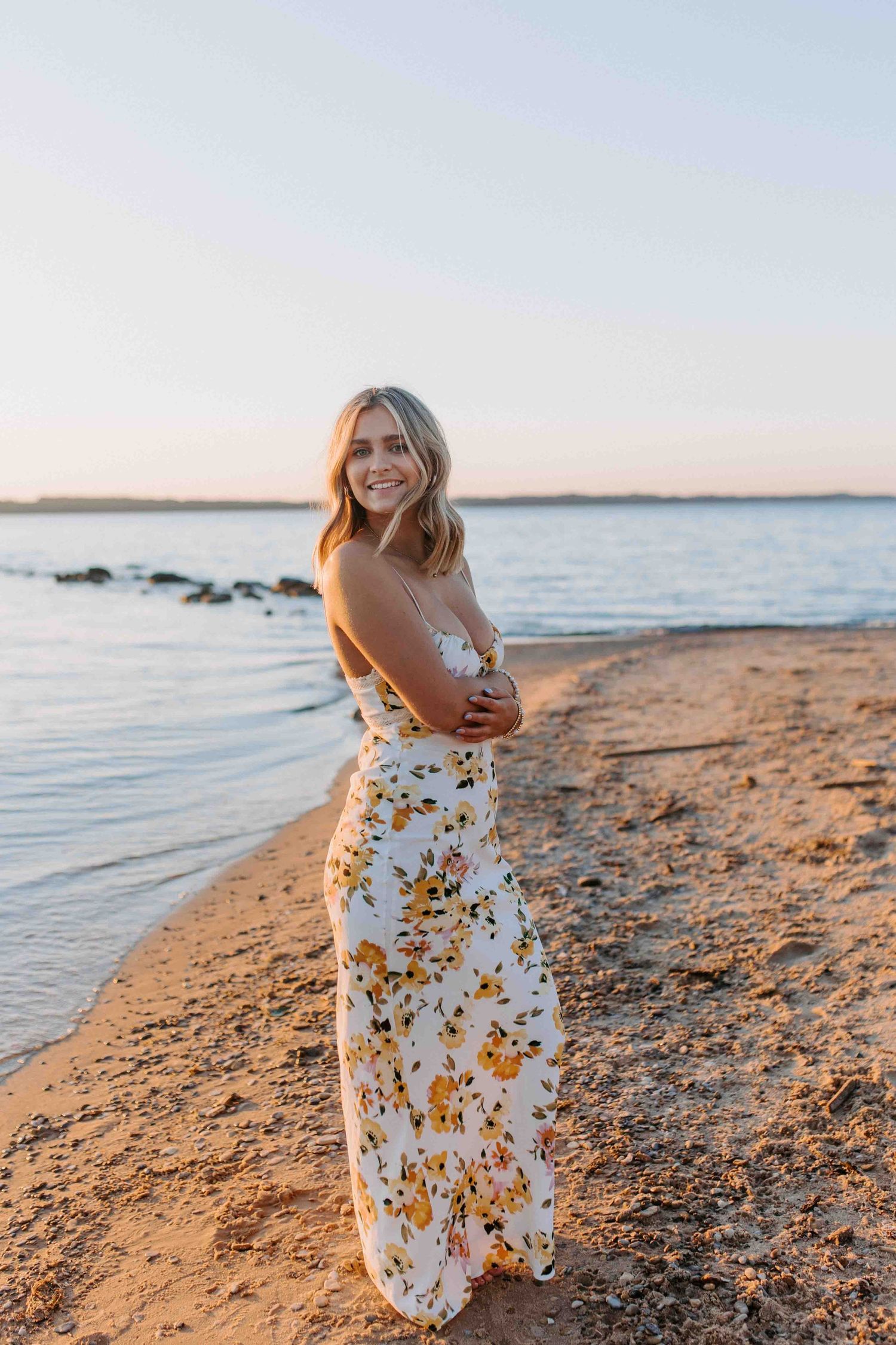 Woman in white floral dress poses casually on sandy beach during sunset photoshoot.