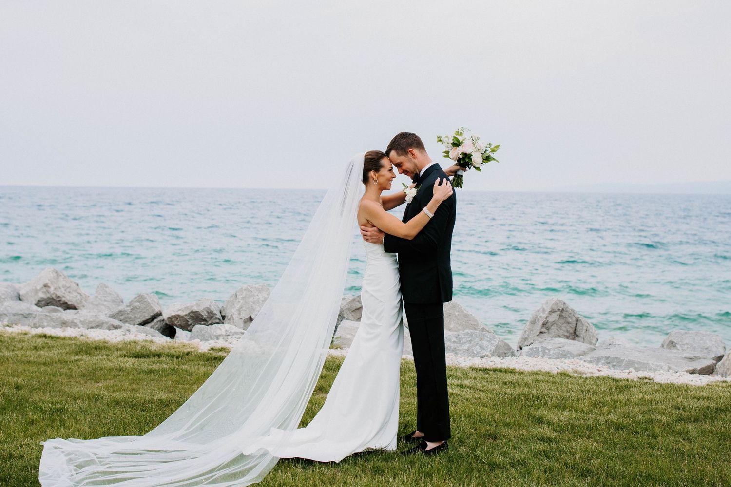Wedding couple embracing on grass overlooking the ocean on their special day.