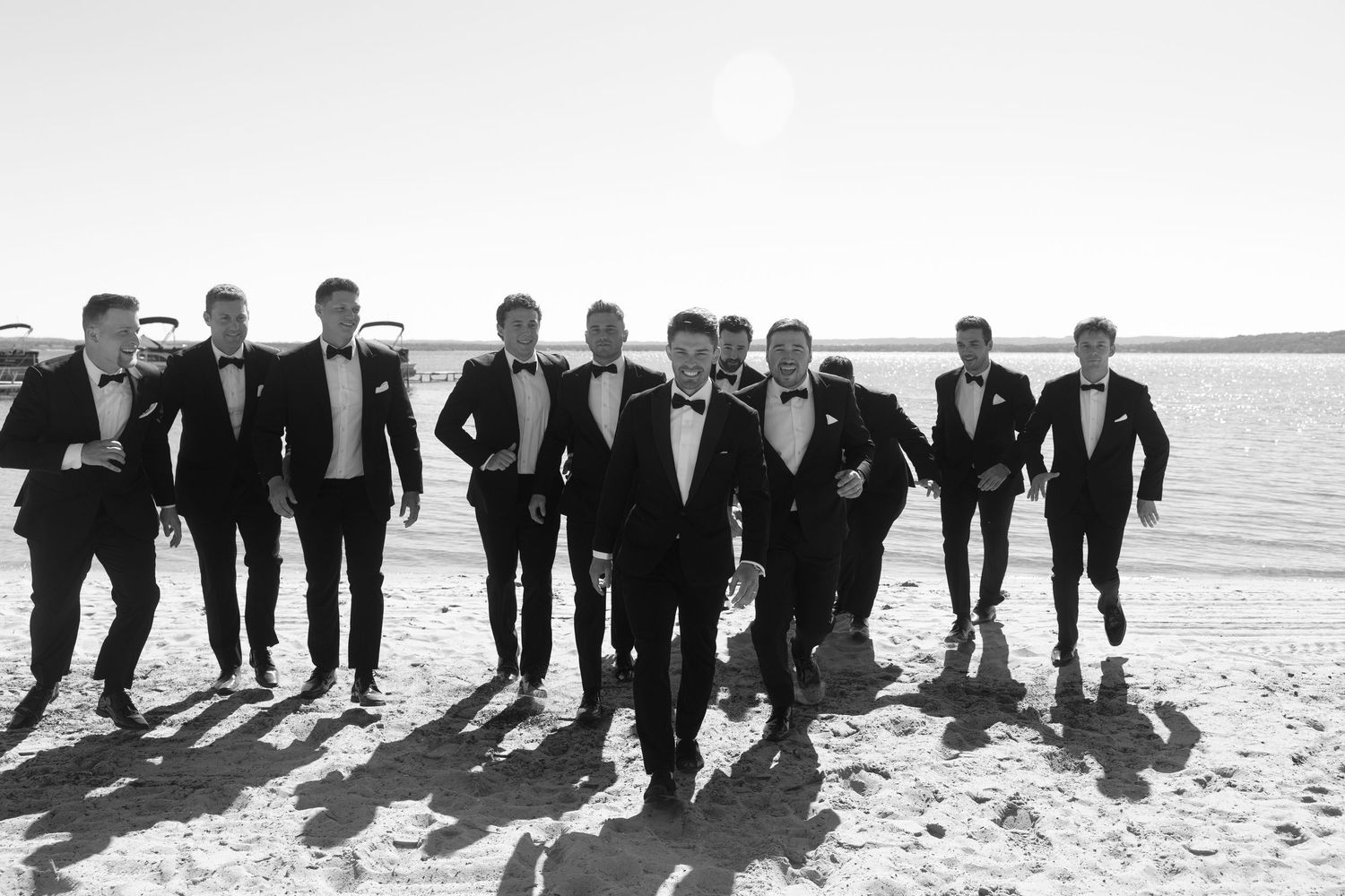 Groomsmen pose together on a beach in formal black attire for a creative wedding photo.
