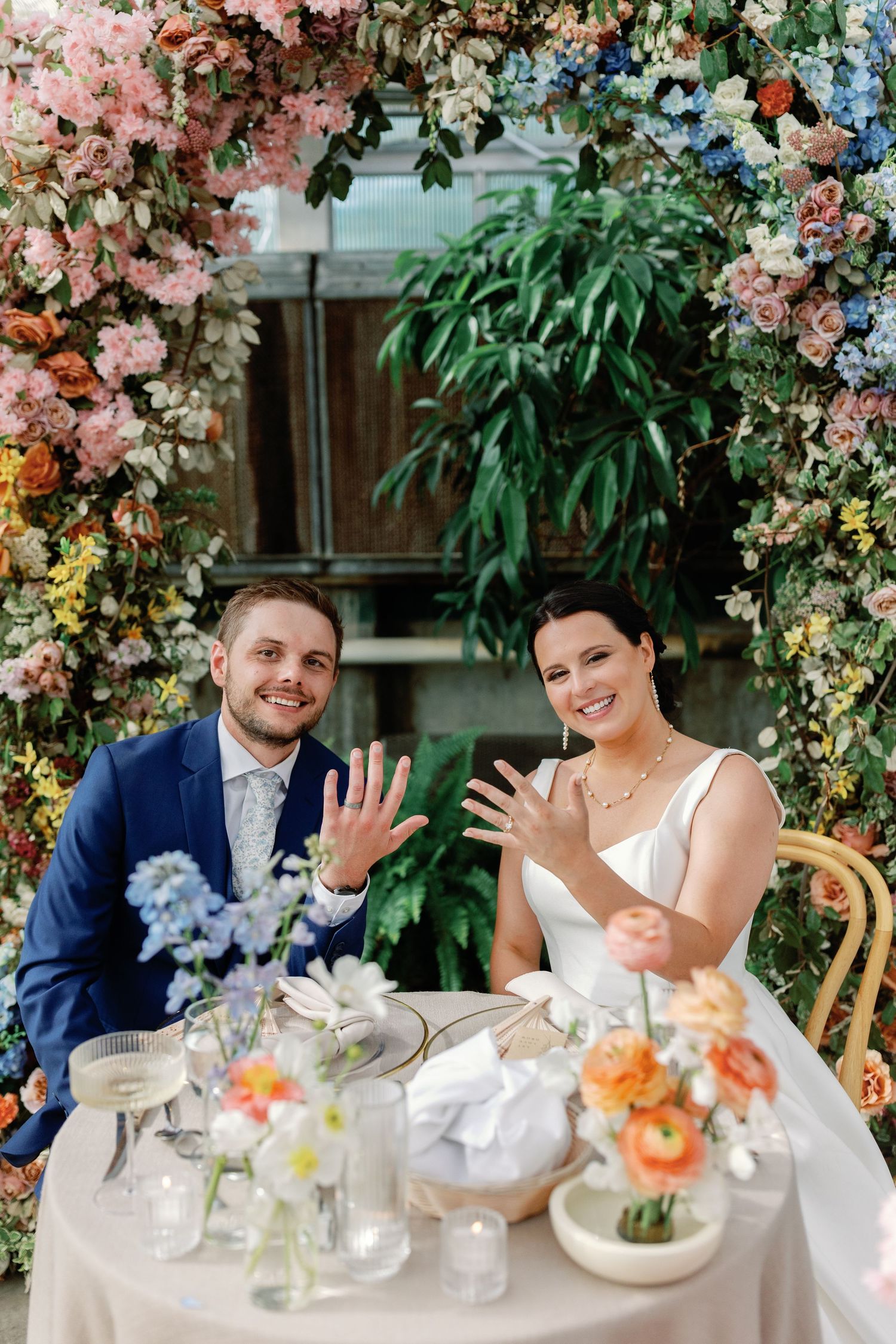 A wedding couple shows off their rings while seated at a table under a floral arch.