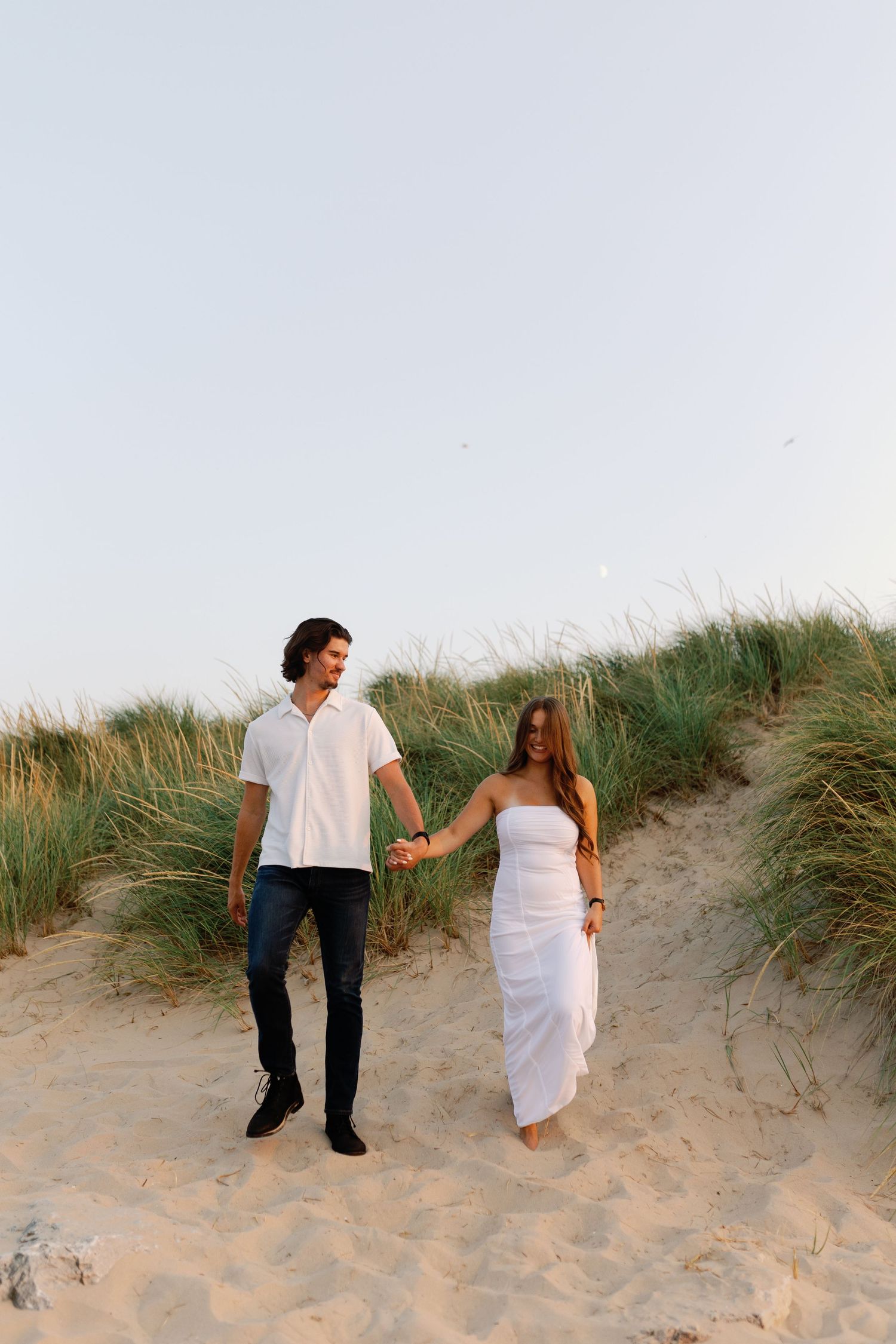 Couple walking together on sandy dunes with beach grass at sunset.