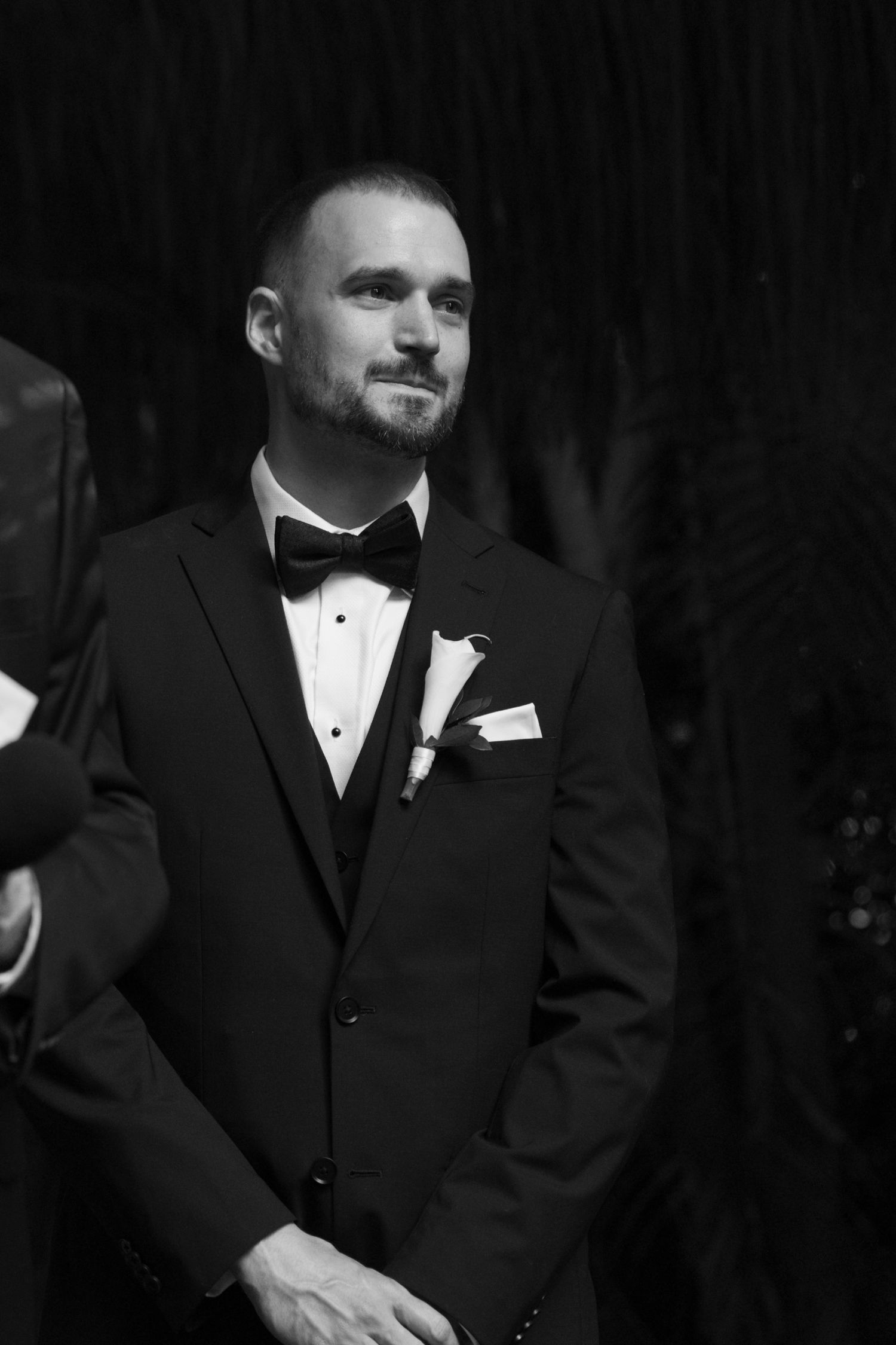 A groom in a classic black tuxedo with bow tie and boutonniere stands during wedding ceremony.