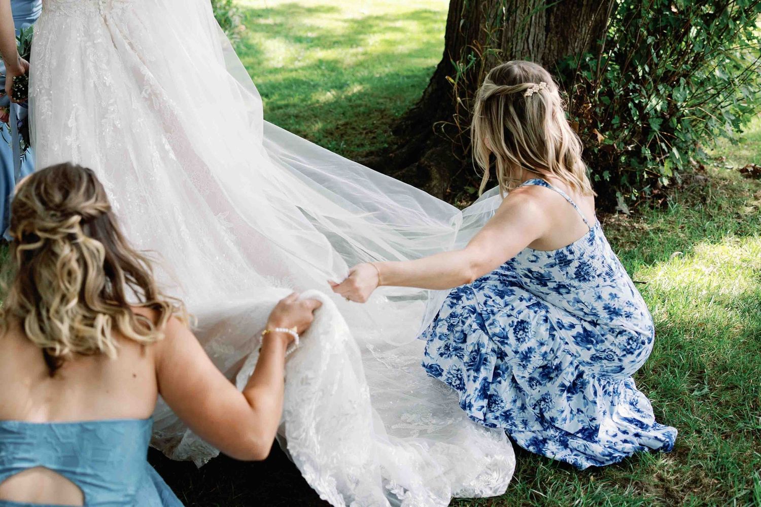 Bridesmaids in blue dresses help arrange the train of a wedding dress outdoors.