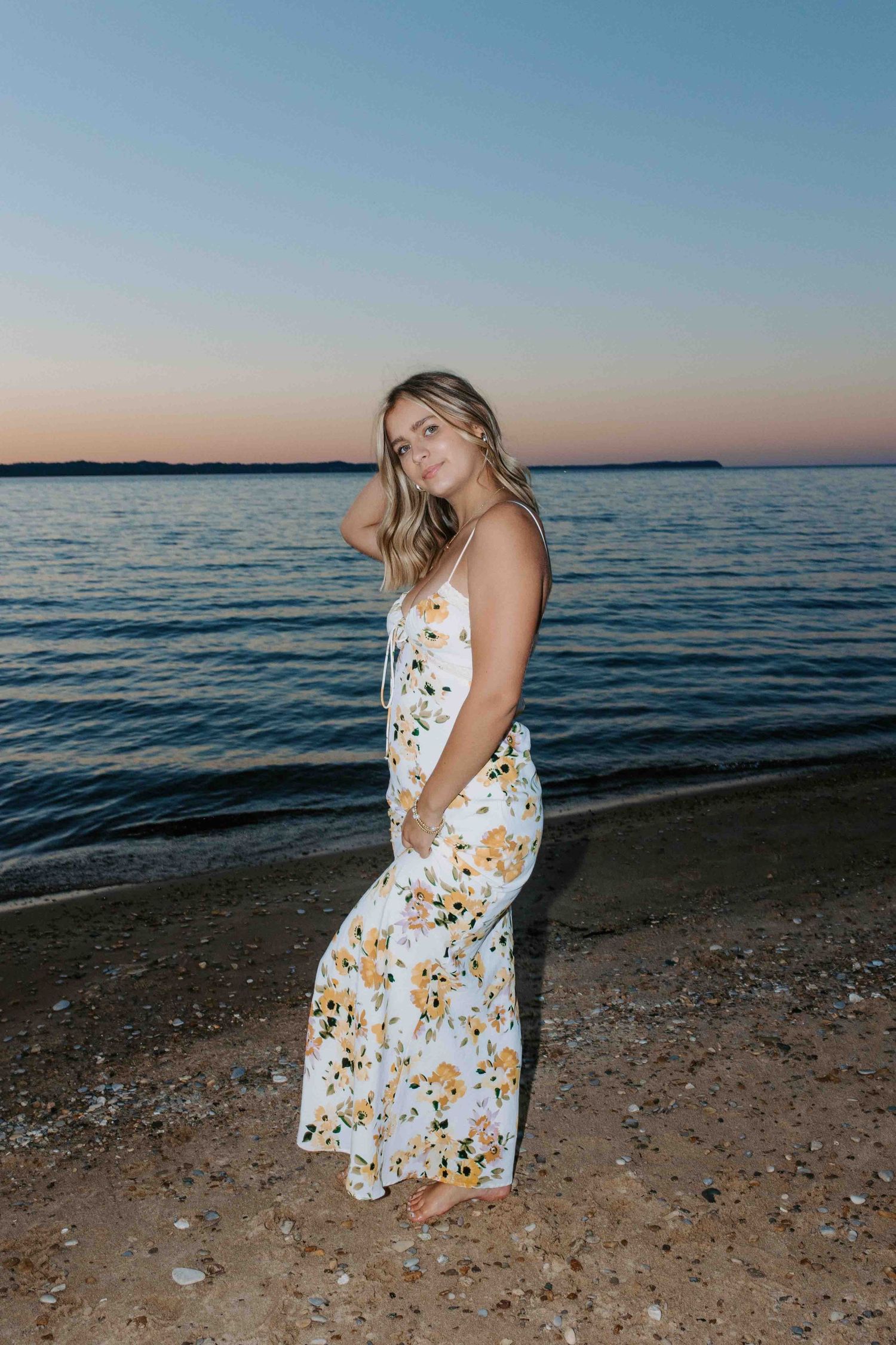 A person poses in a white floral dress on a beach at dusk.