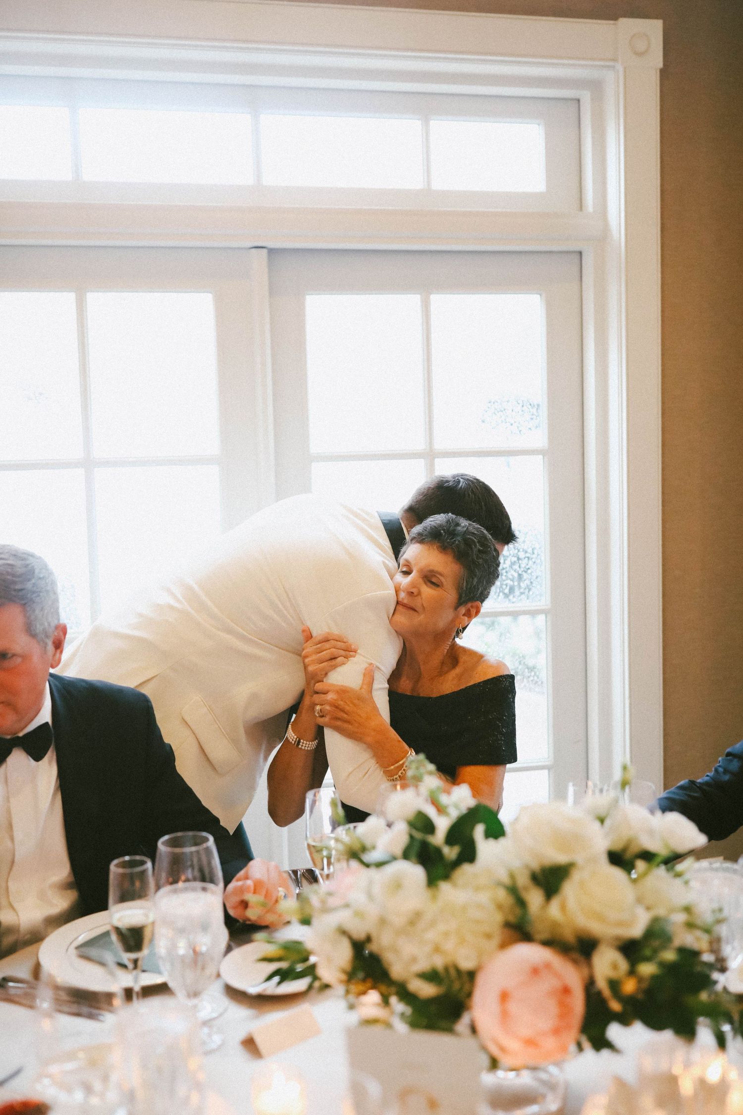 Guests share an intimate moment at an elegant wedding reception with floral centerpieces and wine glasses.
