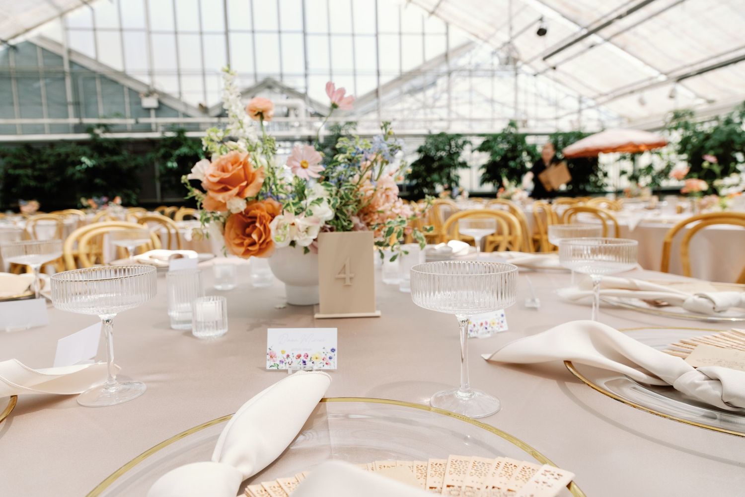 White wedding reception tables set under a clear tent with elegant place settings and gold accents.