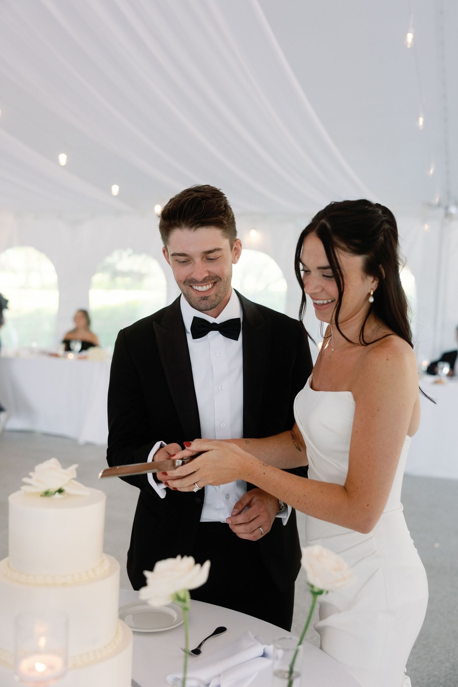 Newlyweds share cake cutting moment in elegant white tent reception.