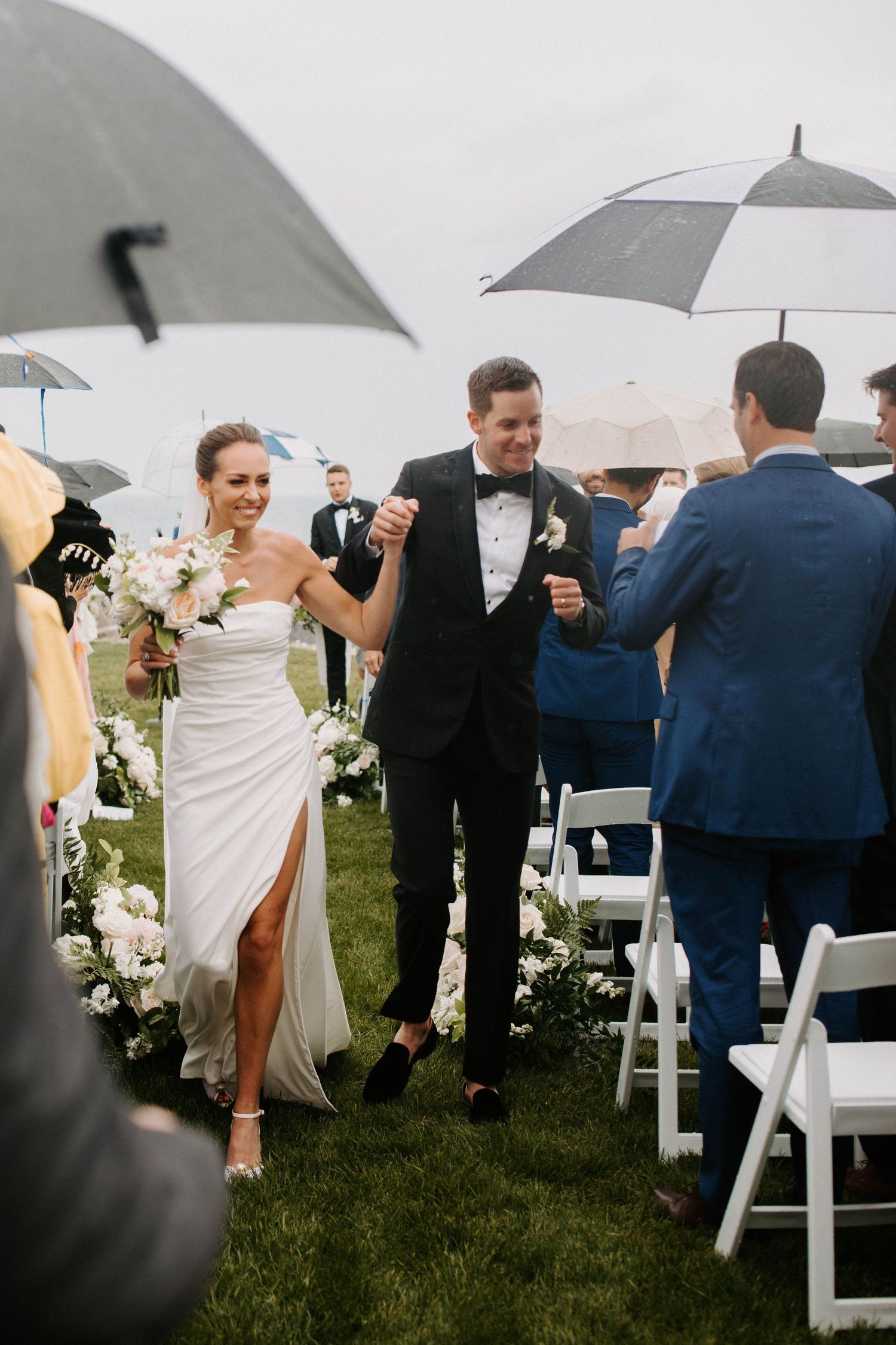 Newlyweds exit ceremony under umbrellas on rainy wedding day.