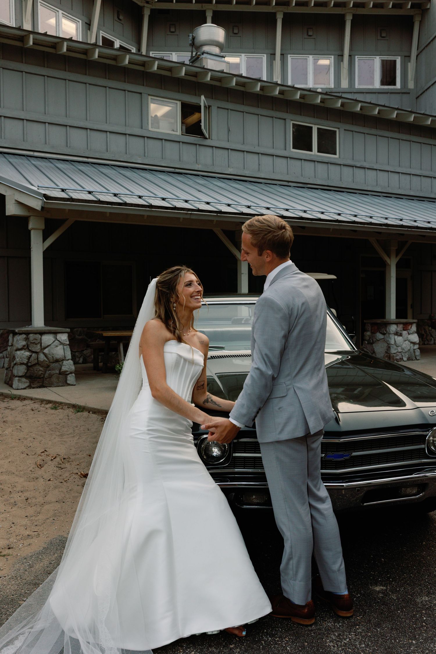 Couple in wedding attire sharing a moment outside a building with vintage cars in background.