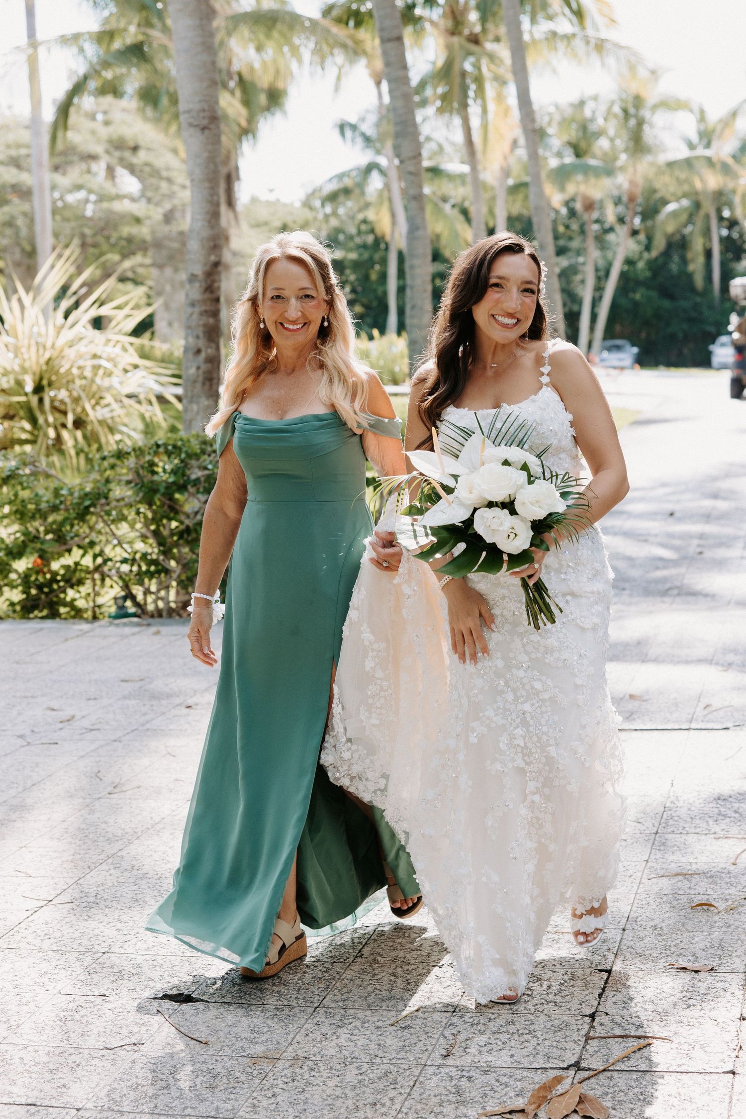 Two women in formal dresses walk along a palm-lined path at a tropical wedding venue.