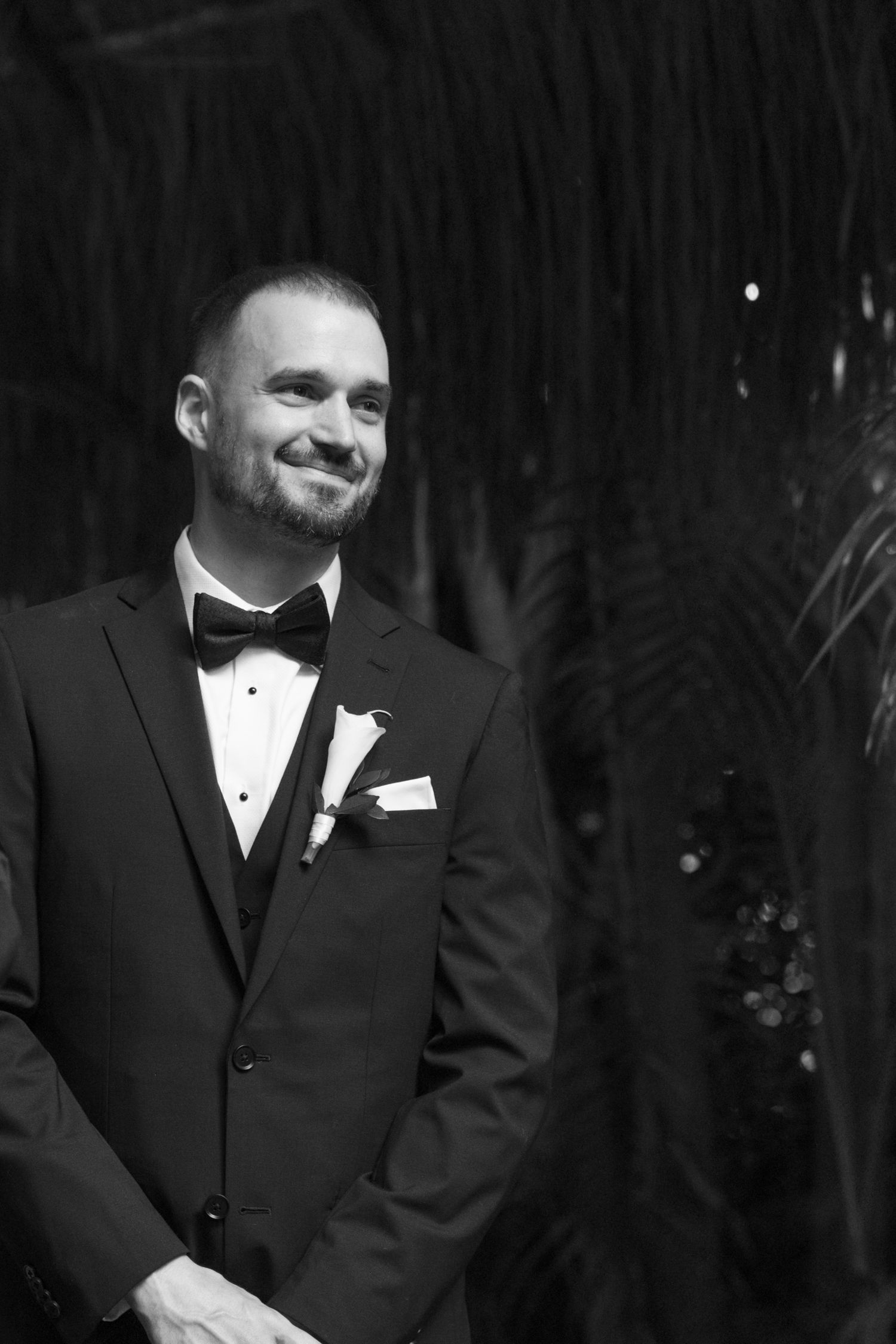 Groom in classic black tuxedo smiling during outdoor wedding ceremony.