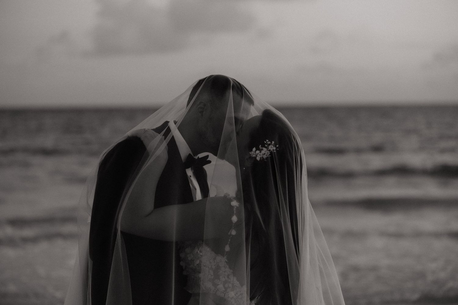 A dramatic black and white beach portrait with flowing veil against the ocean horizon.