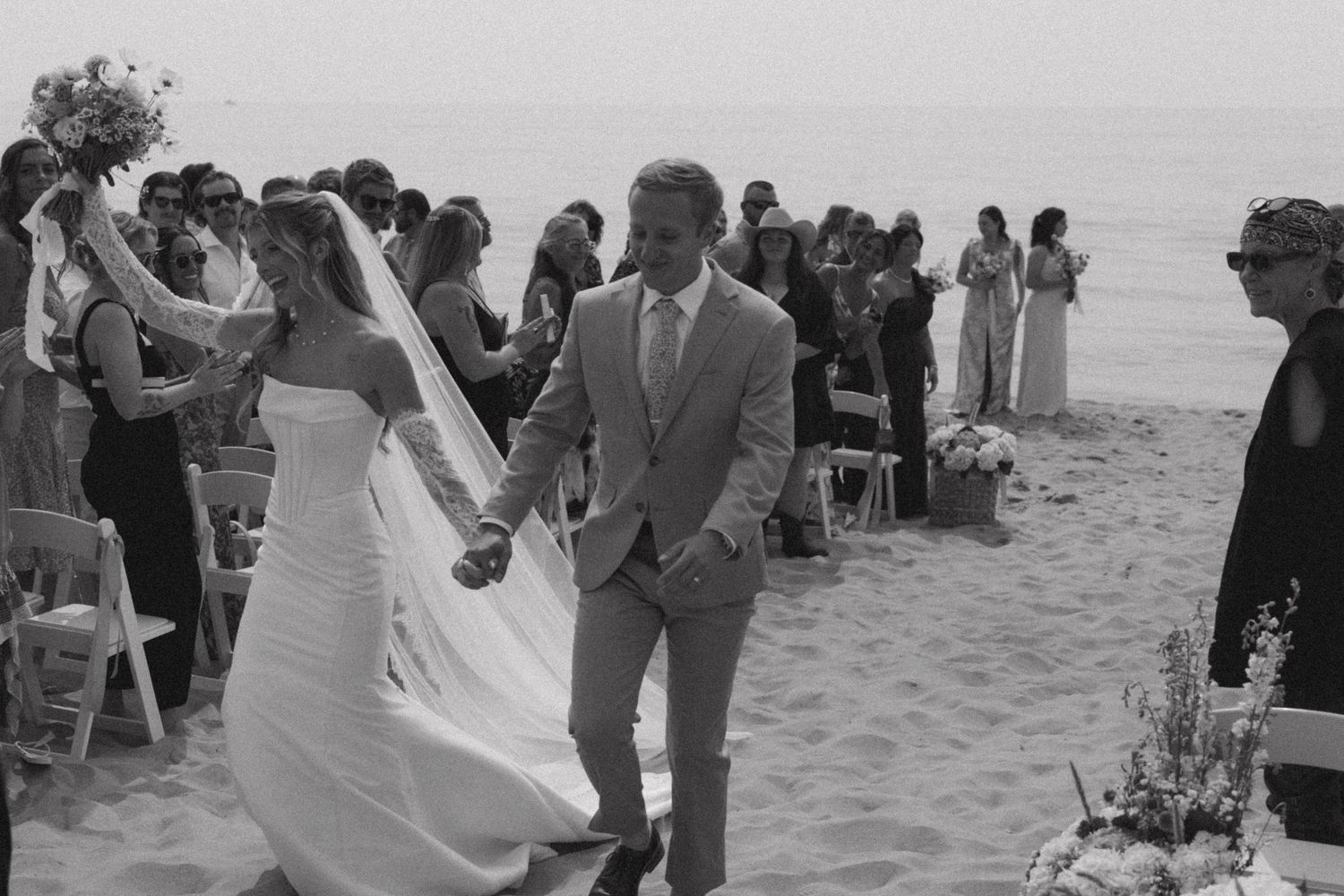 Black and white photo of a beach wedding ceremony with guests seated on white chairs.