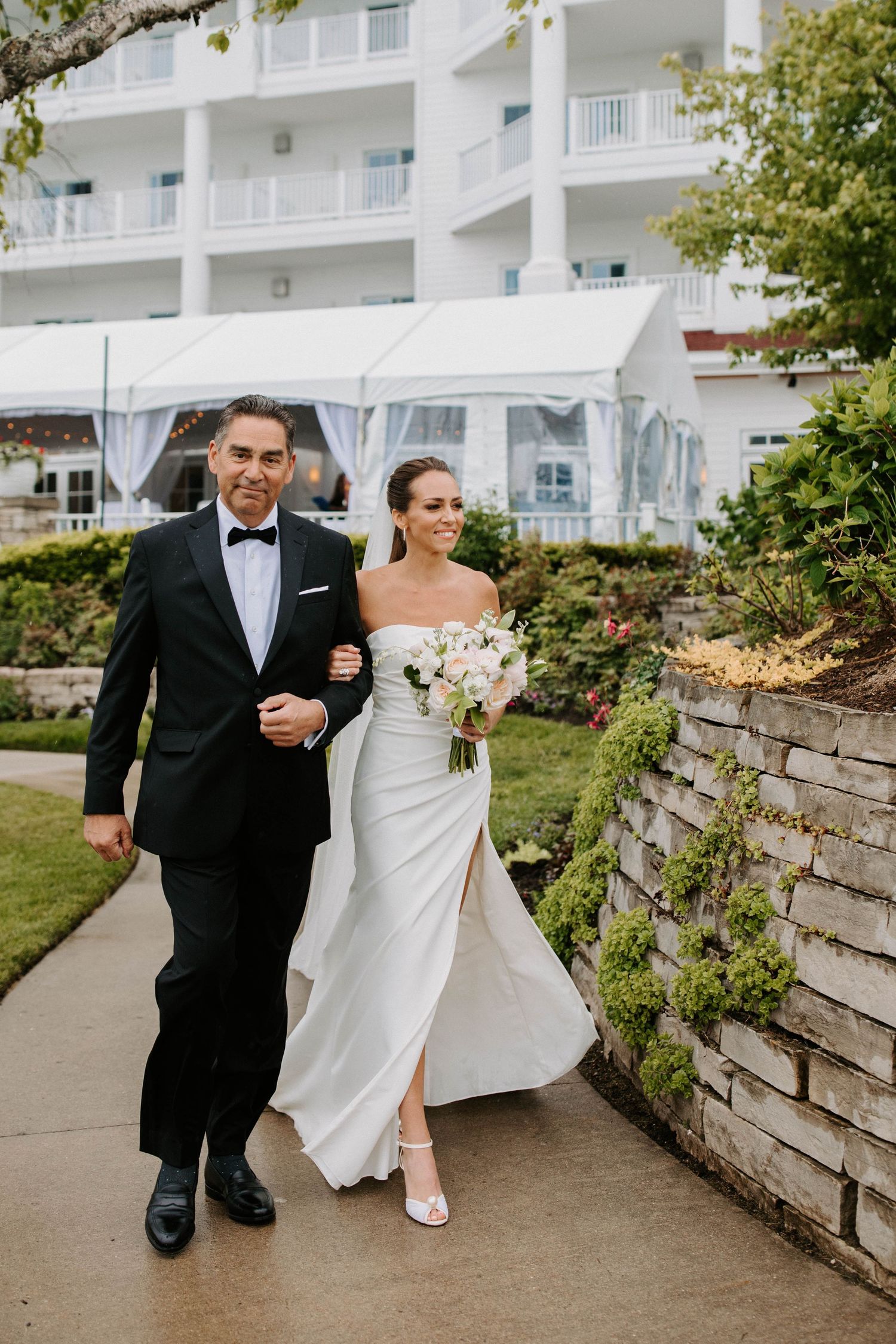 A couple walks along a stone wall pathway outside a white hotel building on their wedding day.
