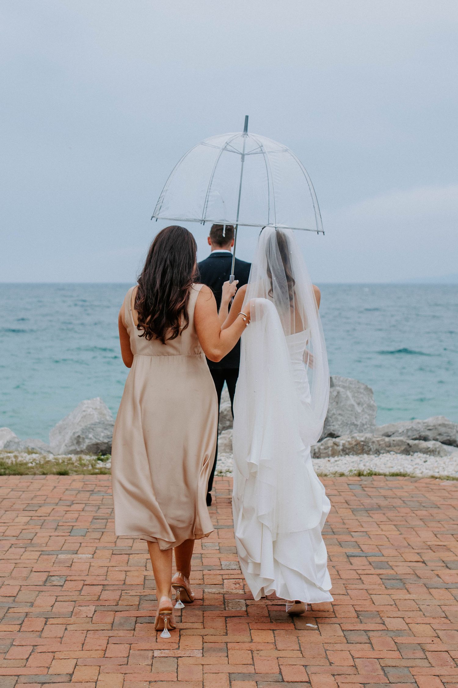 Two figures share an intimate moment under a clear umbrella by the ocean on a brick pathway.