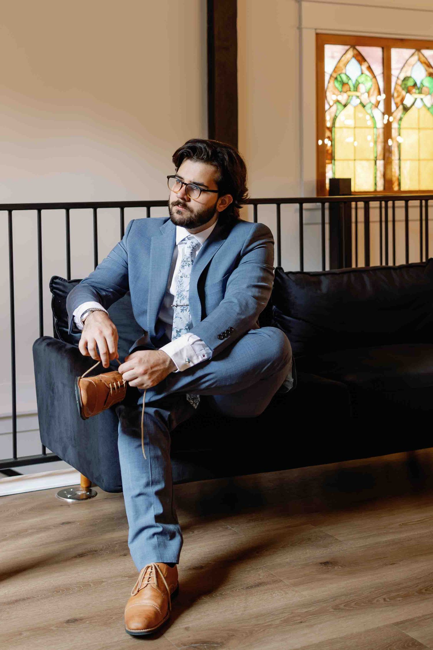 A well-dressed person in a blue suit and glasses sits contemplatively on a black bench near a decorative window.