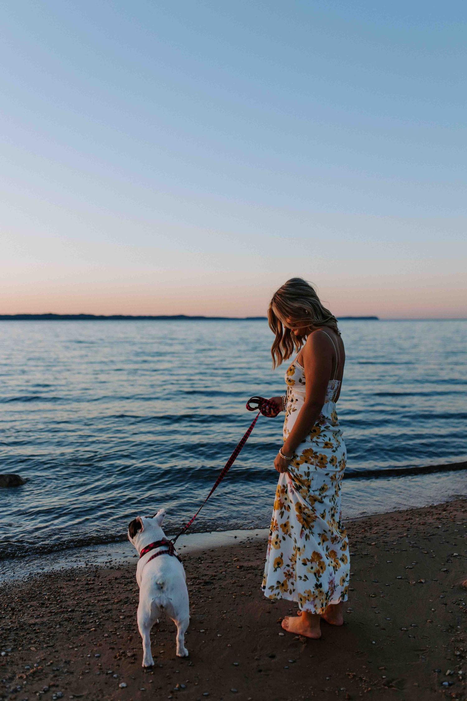 Woman in floral dress walks dog along beach shore during sunset.