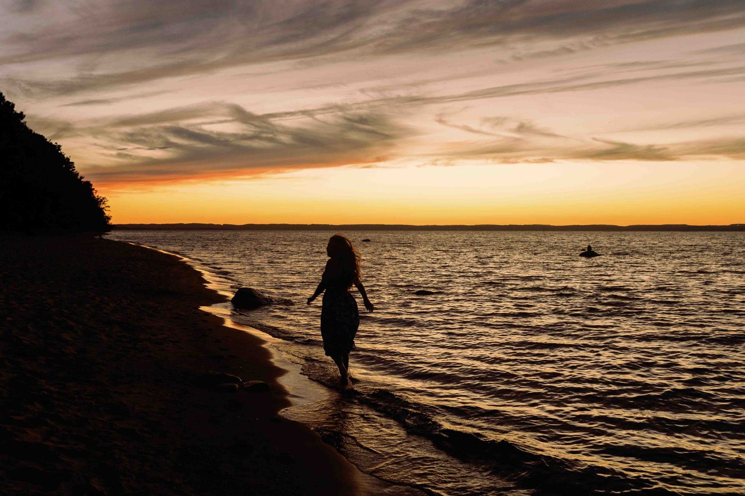 Dark silhouettes walking along beach shoreline at sunset with orange sky.