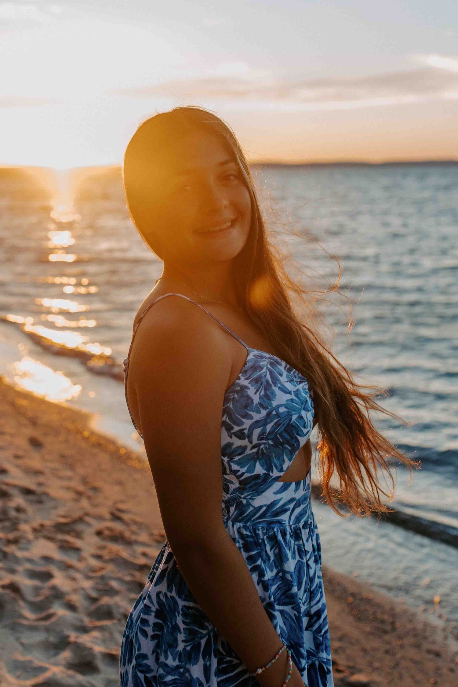 Figure in blue dress stands against dramatic beach sunset with hair blowing in wind.