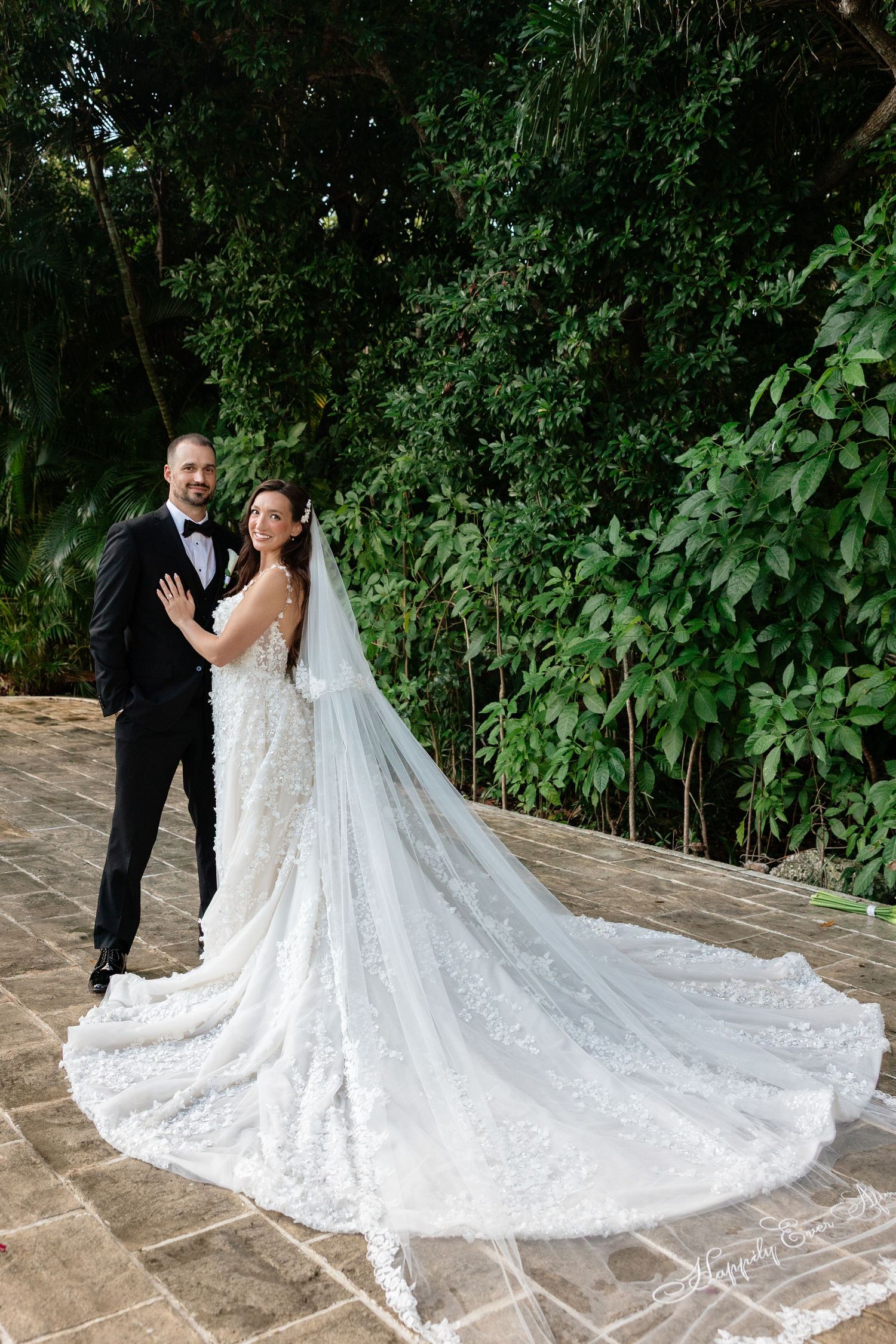 Elegant bridal couple poses with dramatic wedding dress train against garden backdrop.