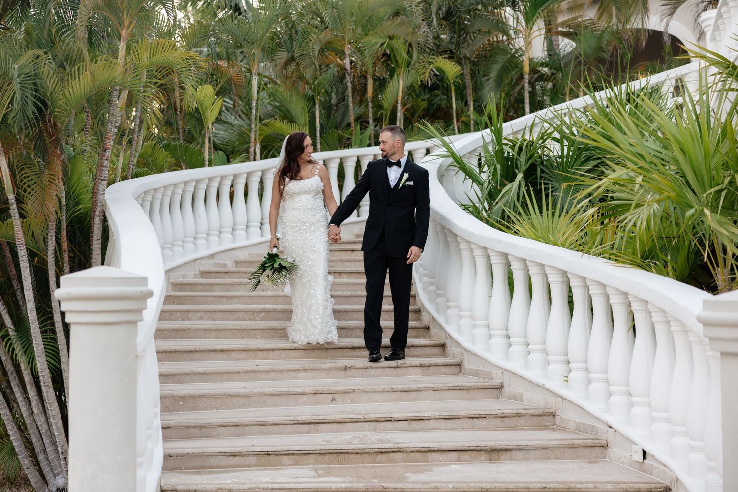 A couple descends an elegant white balustrade staircase surrounded by tropical palm trees at a luxury venue.