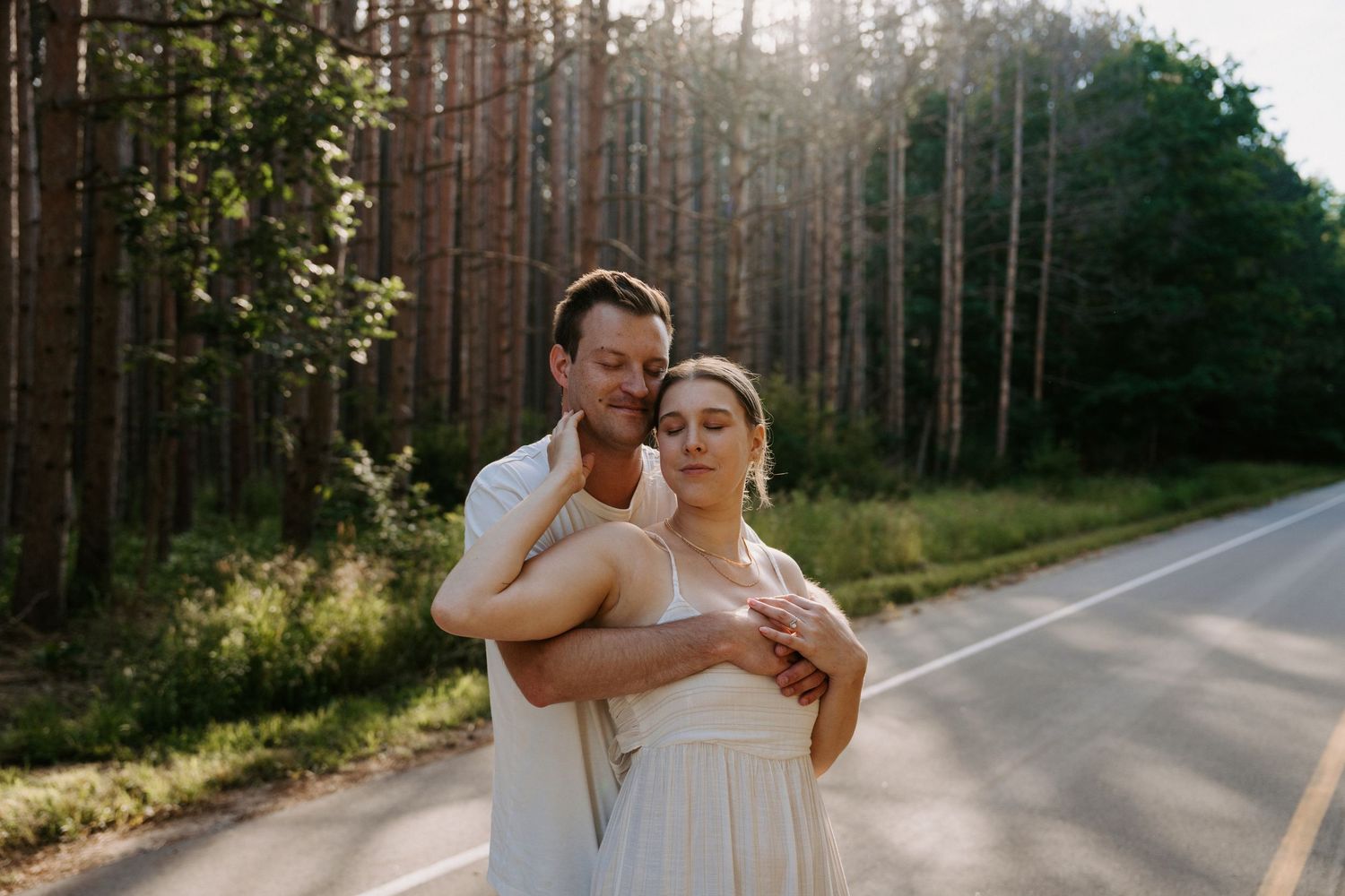 A couple embraces while dancing on a sunlit road surrounded by tall pine trees.