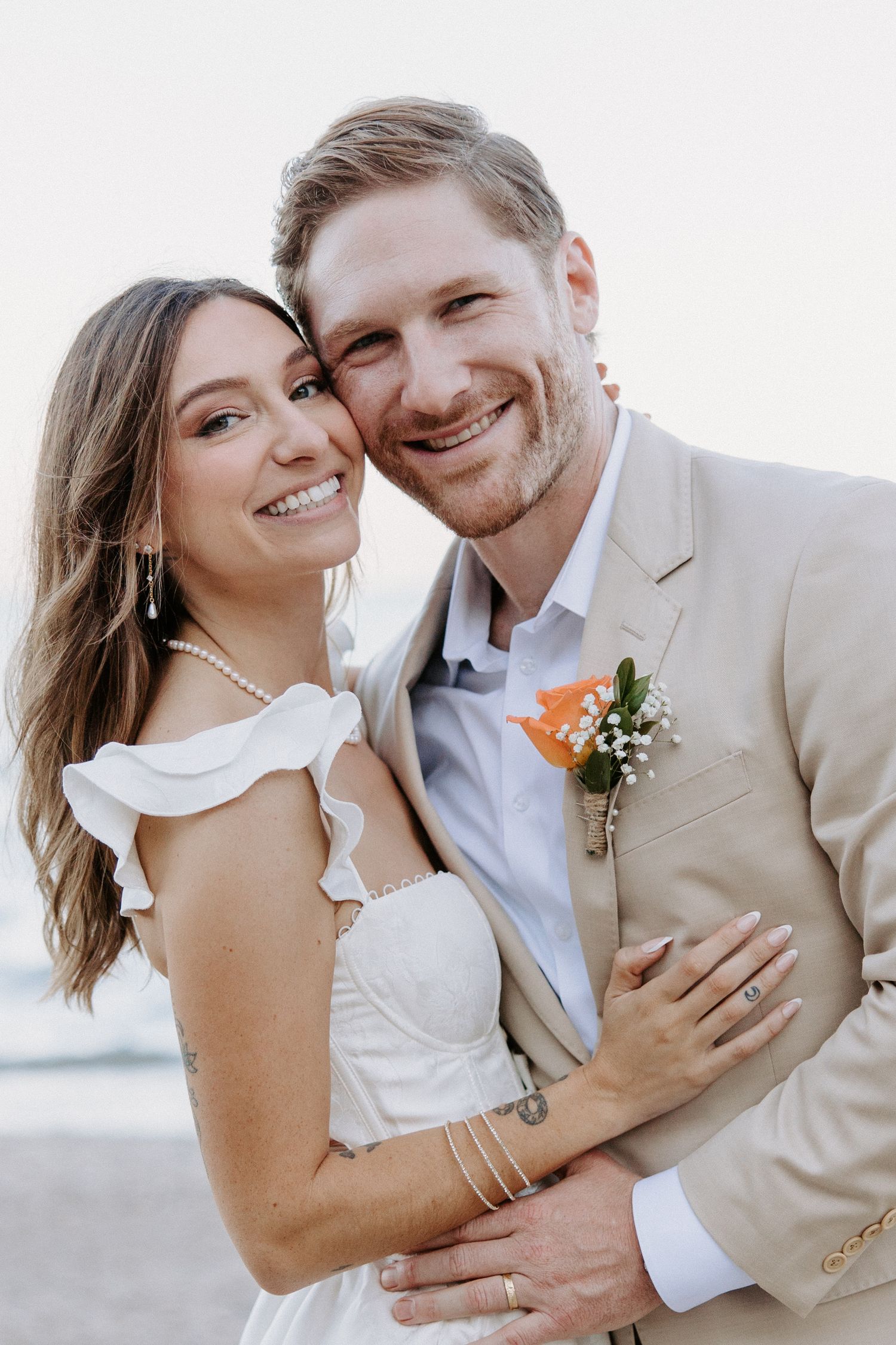 A couple in wedding attire embraces and smiles together in a bright outdoor setting.