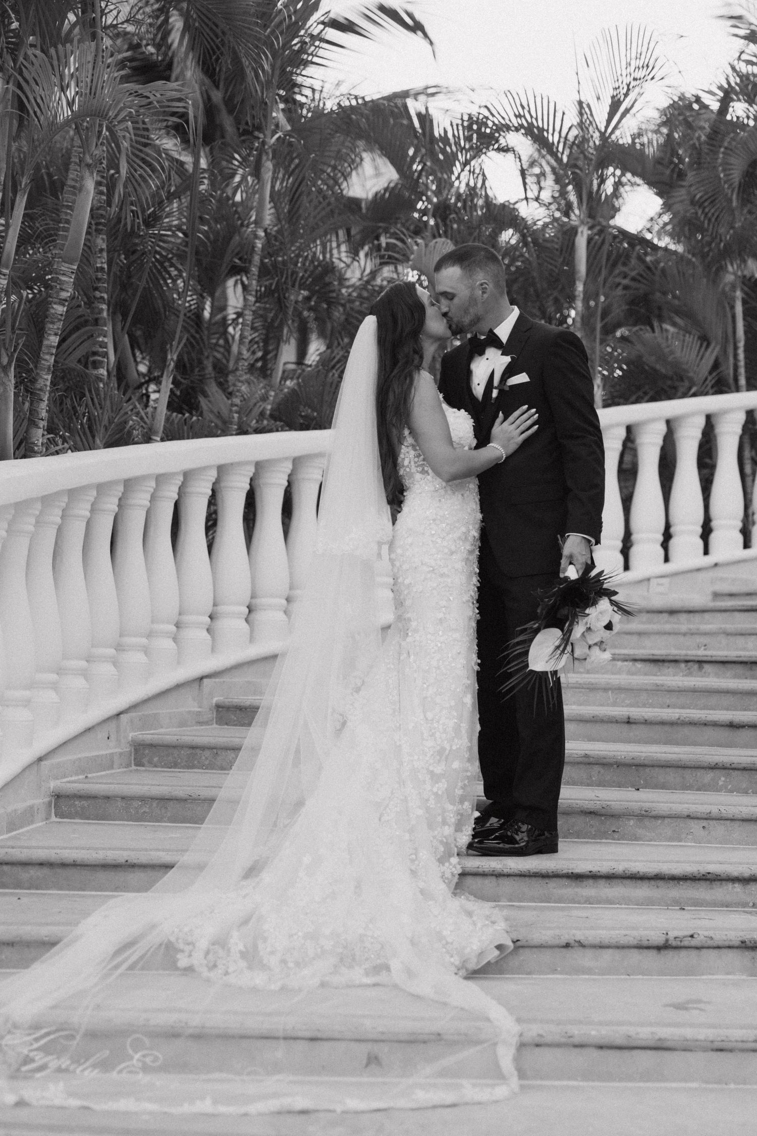 Elegant wedding portrait on white marble stairs surrounded by tropical palm trees.