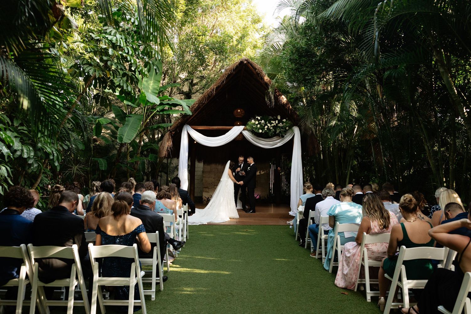 Garden wedding ceremony venue with white chairs arranged in rows under lush green trees.