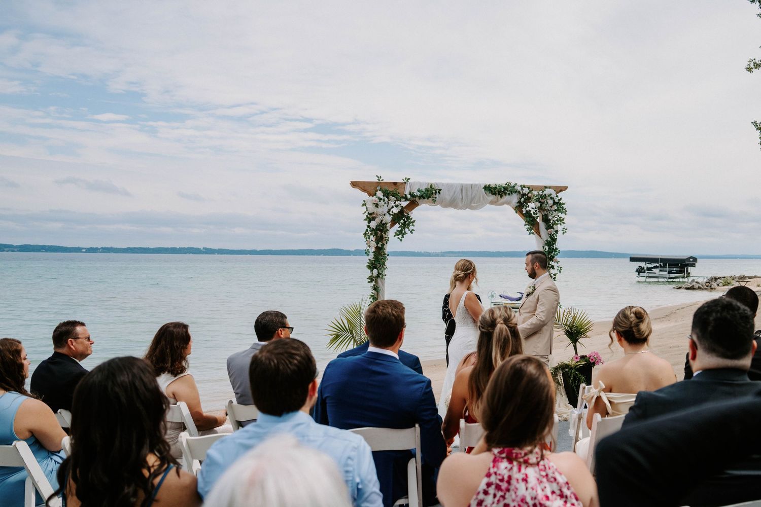 Oceanfront wedding ceremony with floral arch and seated guests against blue water backdrop.