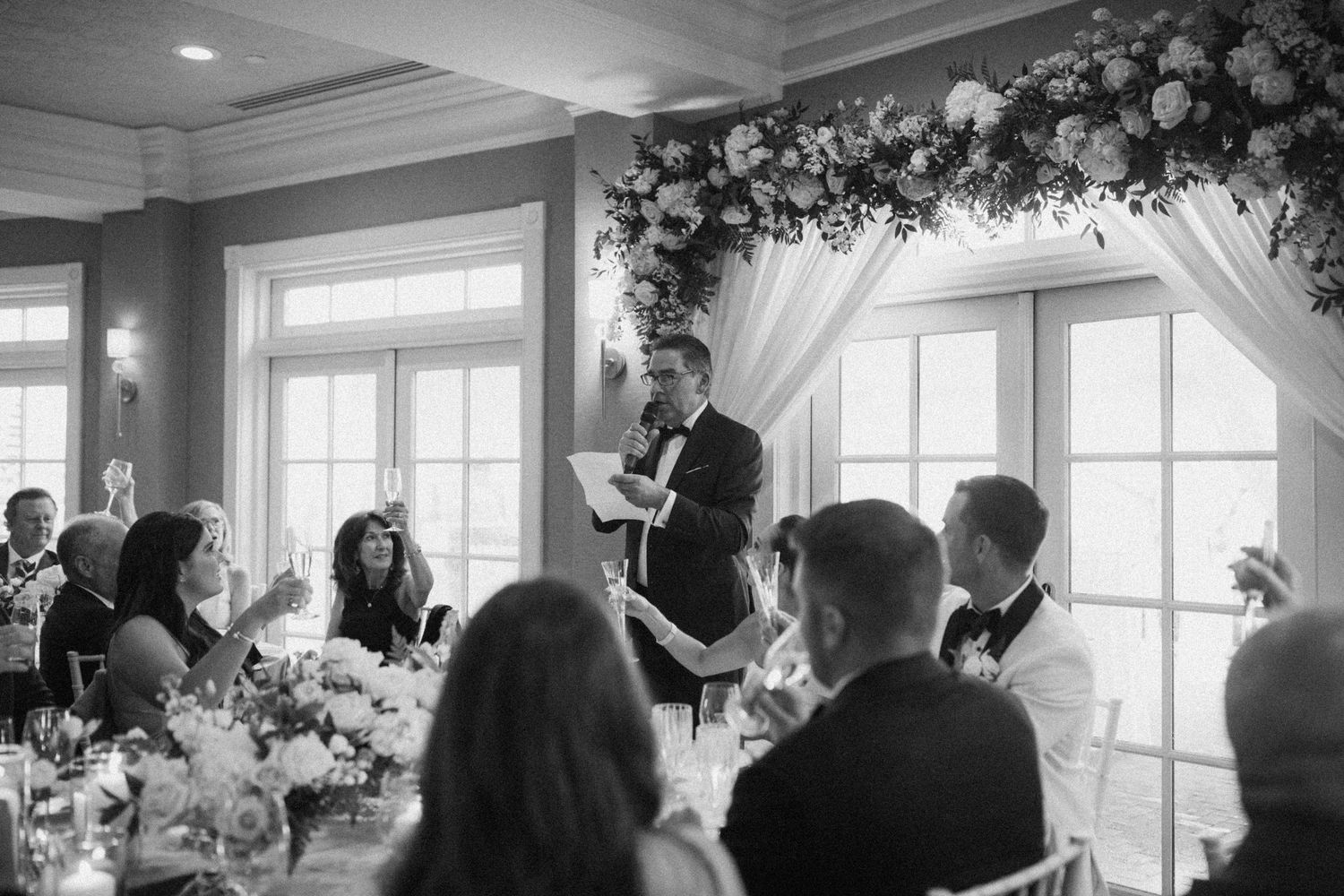 Wedding reception speech under a floral arch with guests seated at decorated tables.