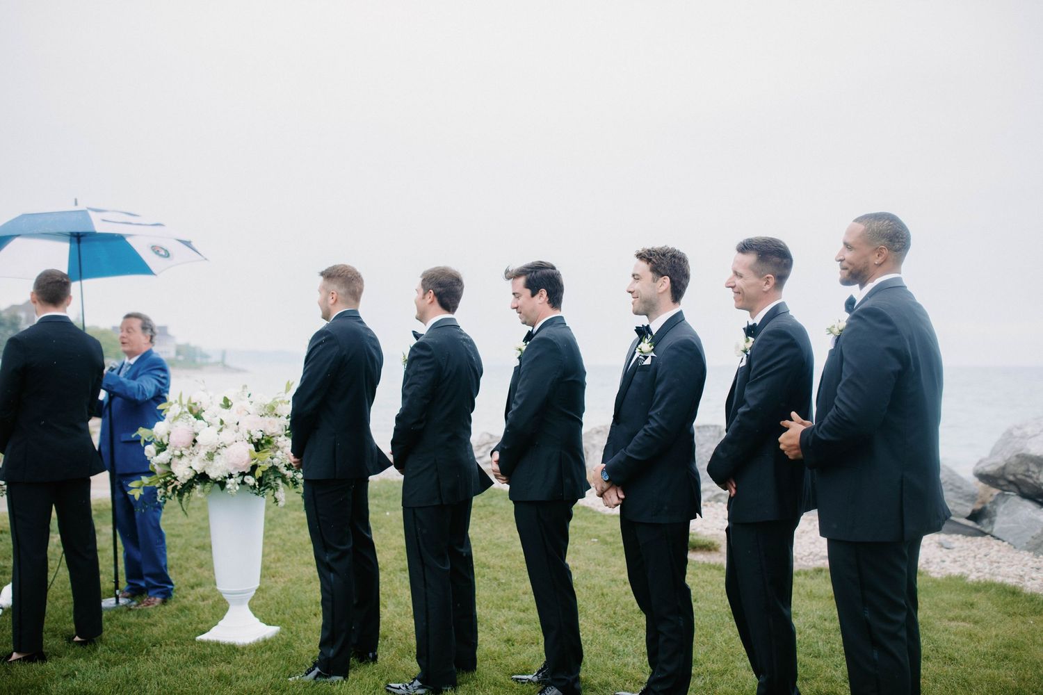 Wedding party members line up on grass wearing formal attire during an outdoor ceremony.