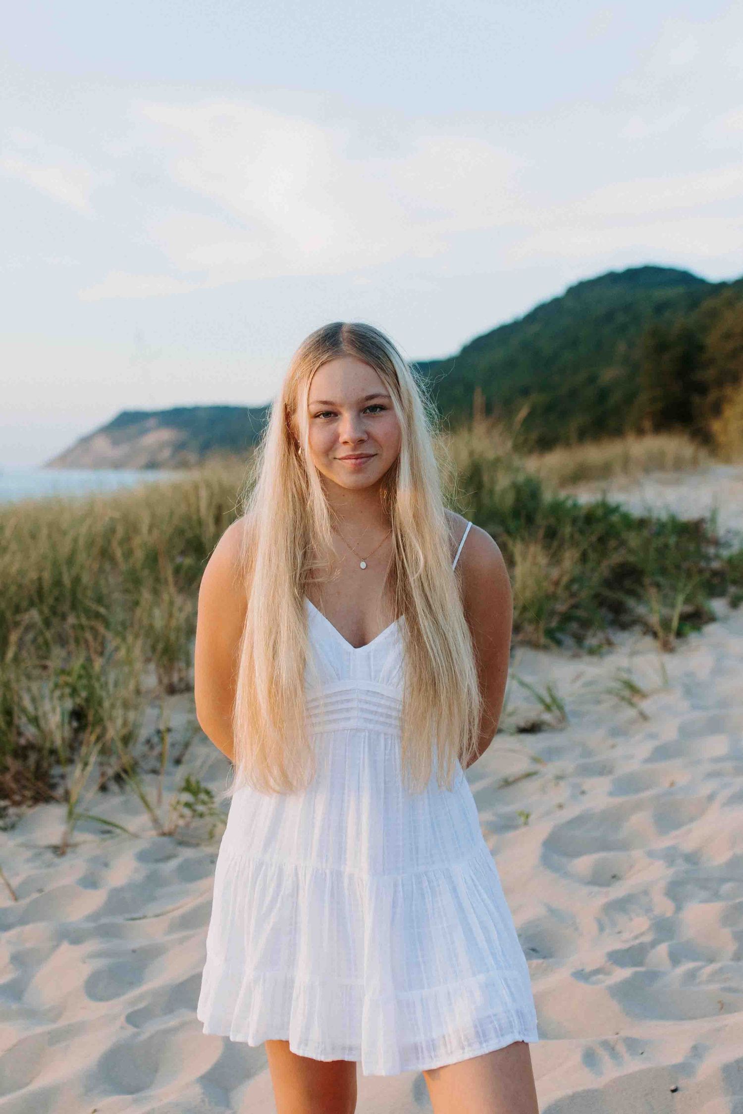 Person in flowing white dress poses on sandy beach with mountains in background.