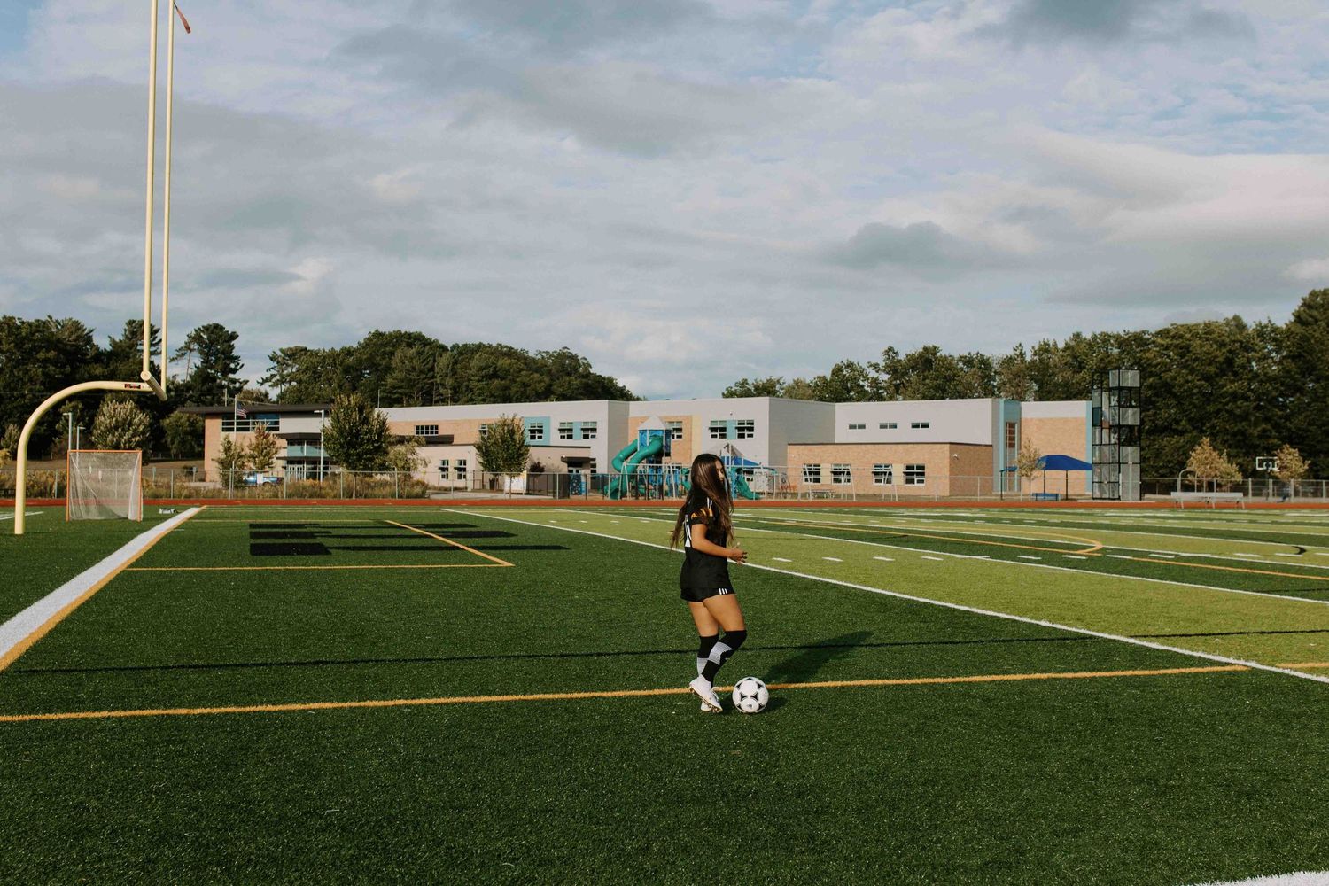 Soccer player practices ball control on empty turf field with school buildings in background.