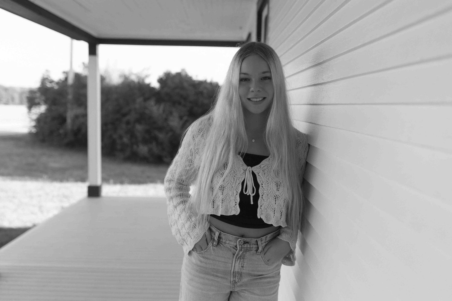 A casual outdoor photo shoot featuring light denim jeans, a white knit sweater and black sneakers on a porch.