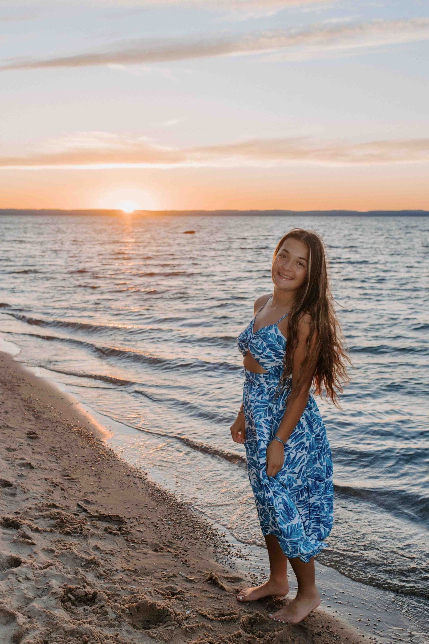 Figure in blue patterned dress poses gracefully on beach at sunset with waves in background.