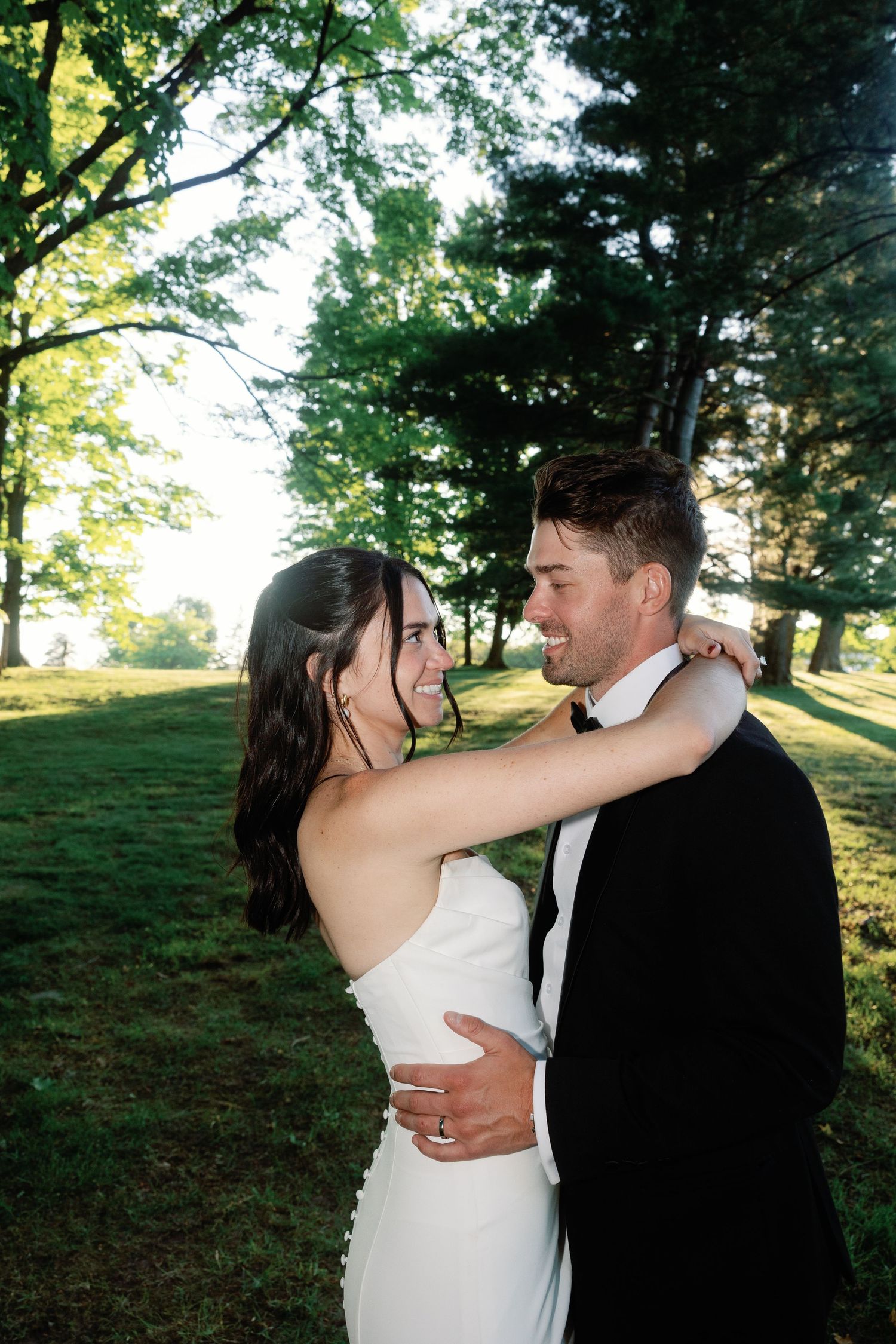 Newlyweds embrace in a sunlit outdoor garden setting with trees in the background.